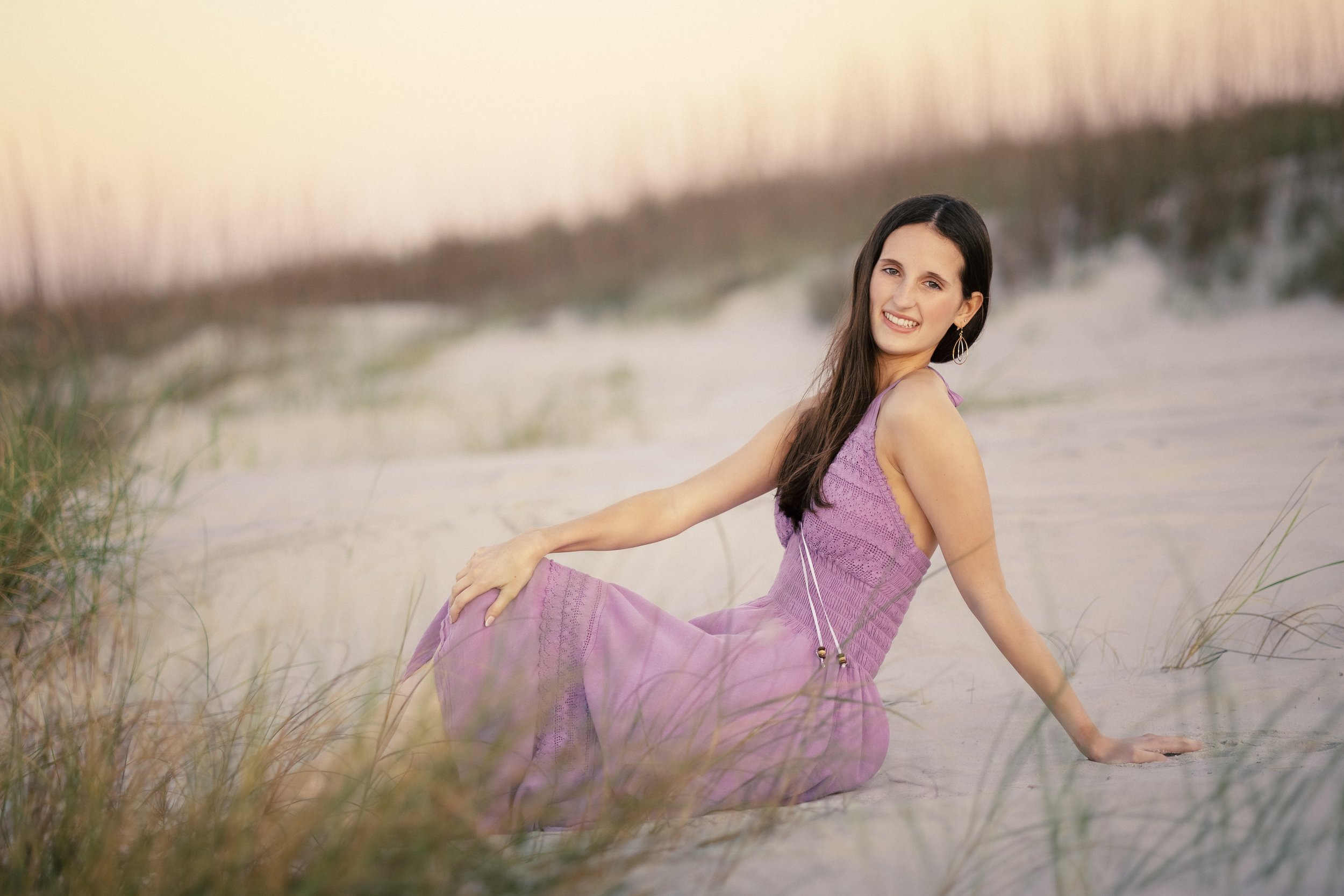 A woman in a purple dress sitting on the sandy beach among grass, smiling at the camera with a sunset sky in the background.