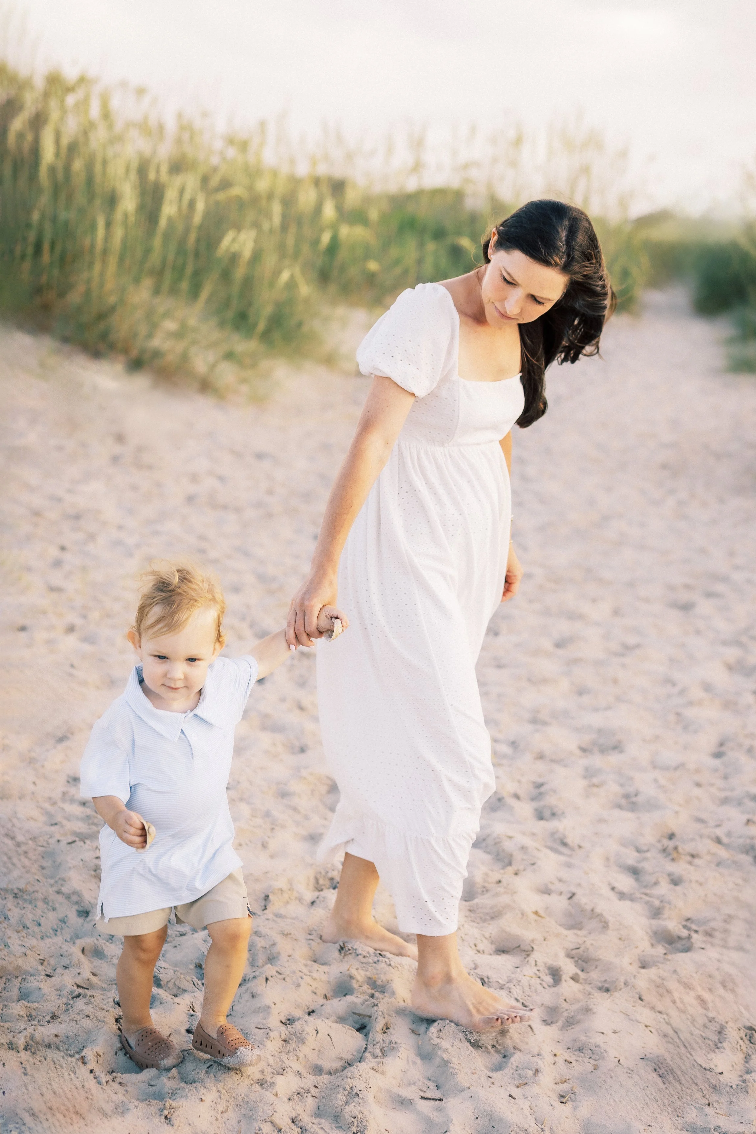 A woman and a young boy walking barefoot on a sandy beach, holding hands, with grassy dunes in the background during sunset or late afternoon.Photographer Amelia Island, Jacksonville, St Augustine, St Simons Island, Savannah photographer