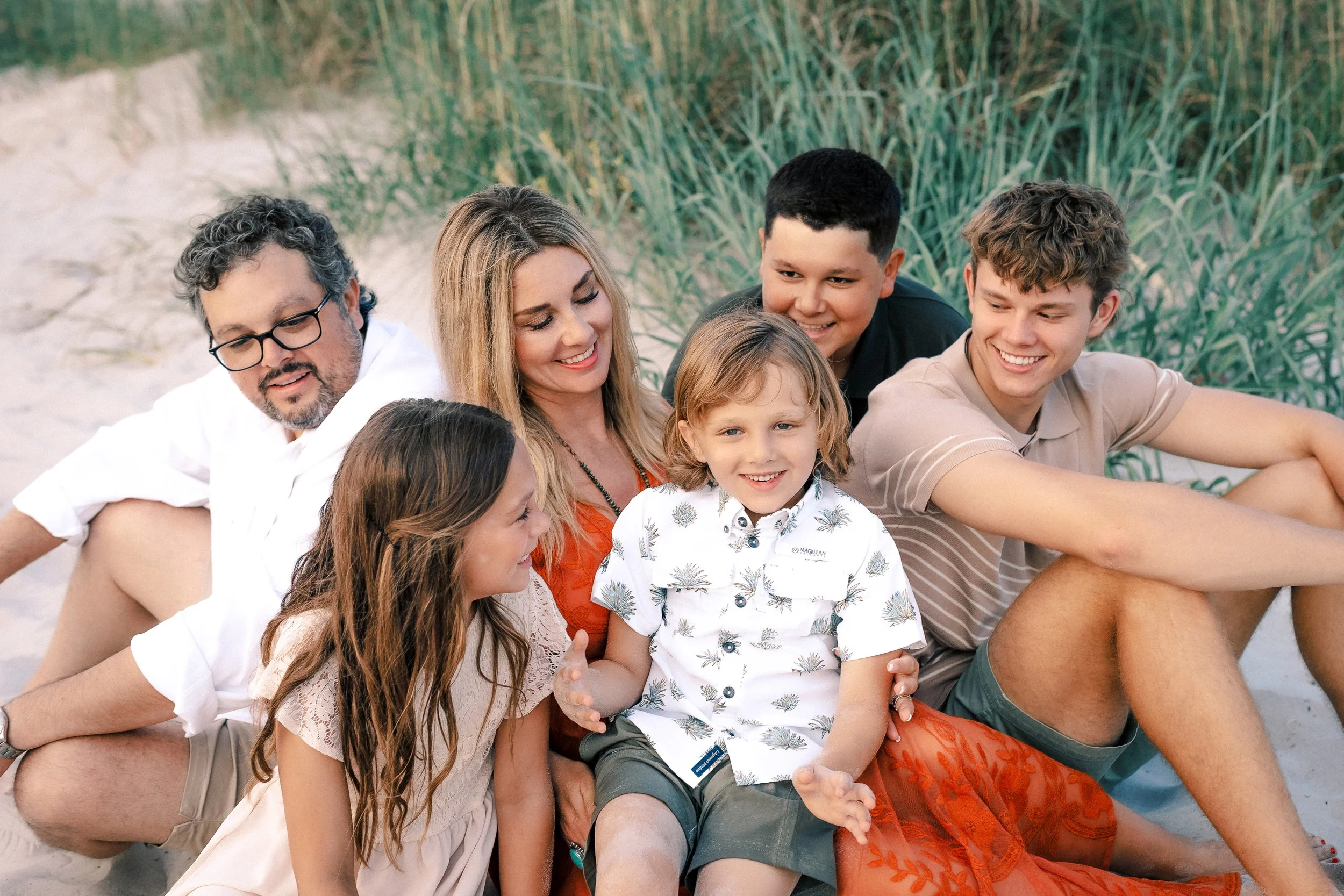 A family of seven people sitting on the beach, smiling and enjoying a sunny day. There are three adults and four children, with greenery in the background.