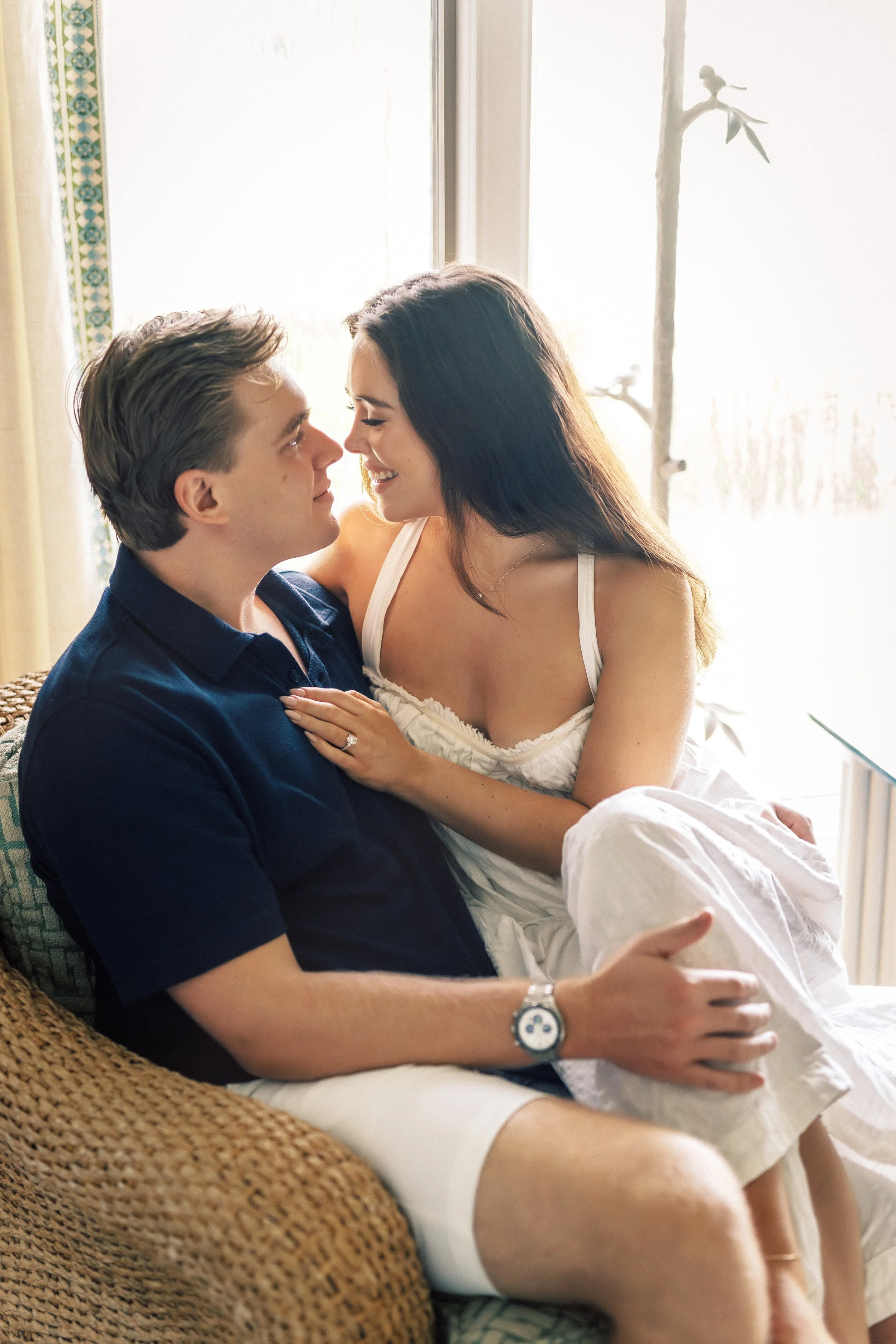 A young couple sitting closely together on a wicker chair near a window, smiling and about to kiss, with bright sunlight behind them.