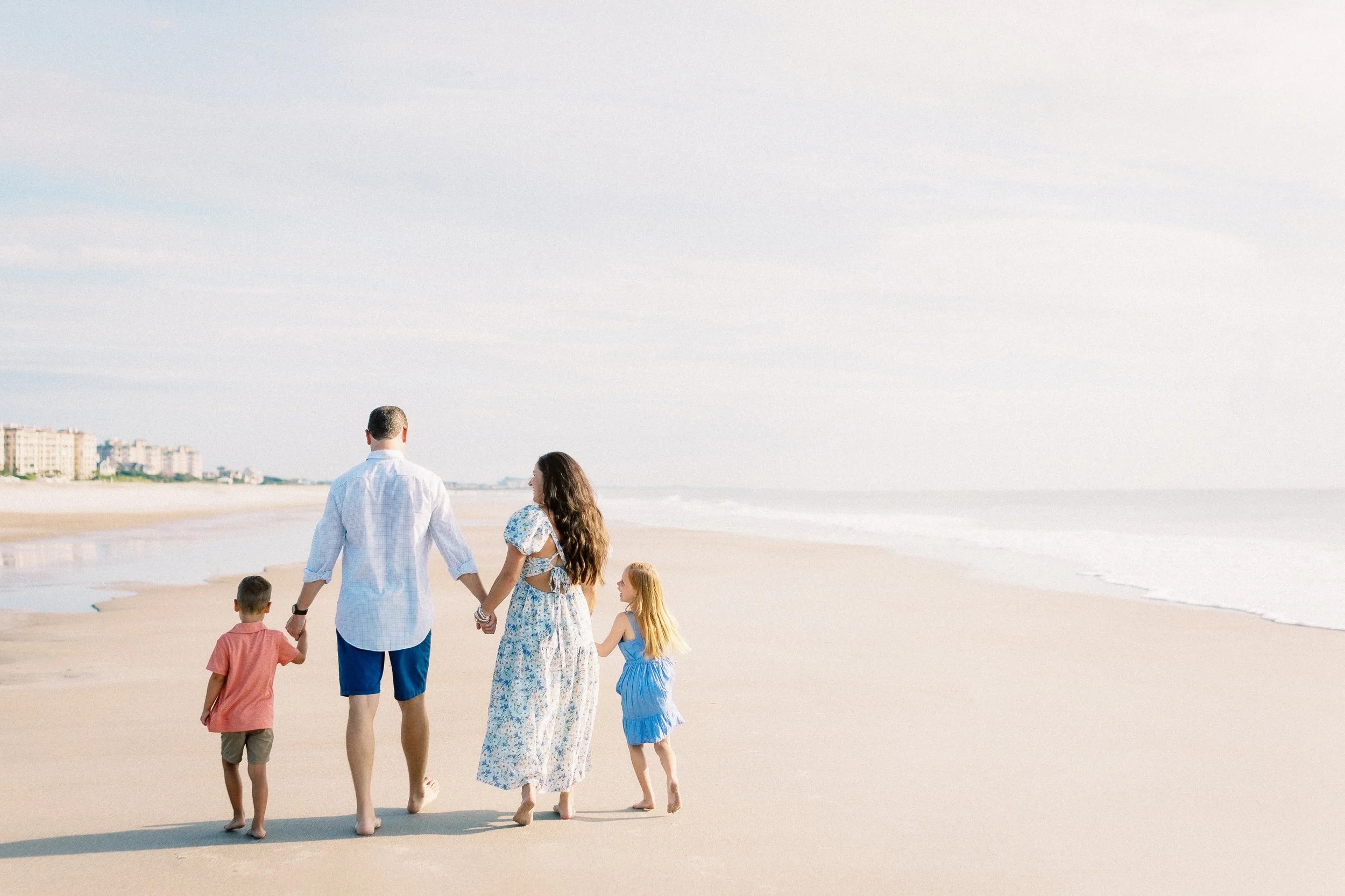 A family of five walking on the beach, holding hands, with the ocean and a distant cityscape in the background.Photographer Amelia Island, Jacksonville, St Augustine, St Simons Island, Savannah photographer