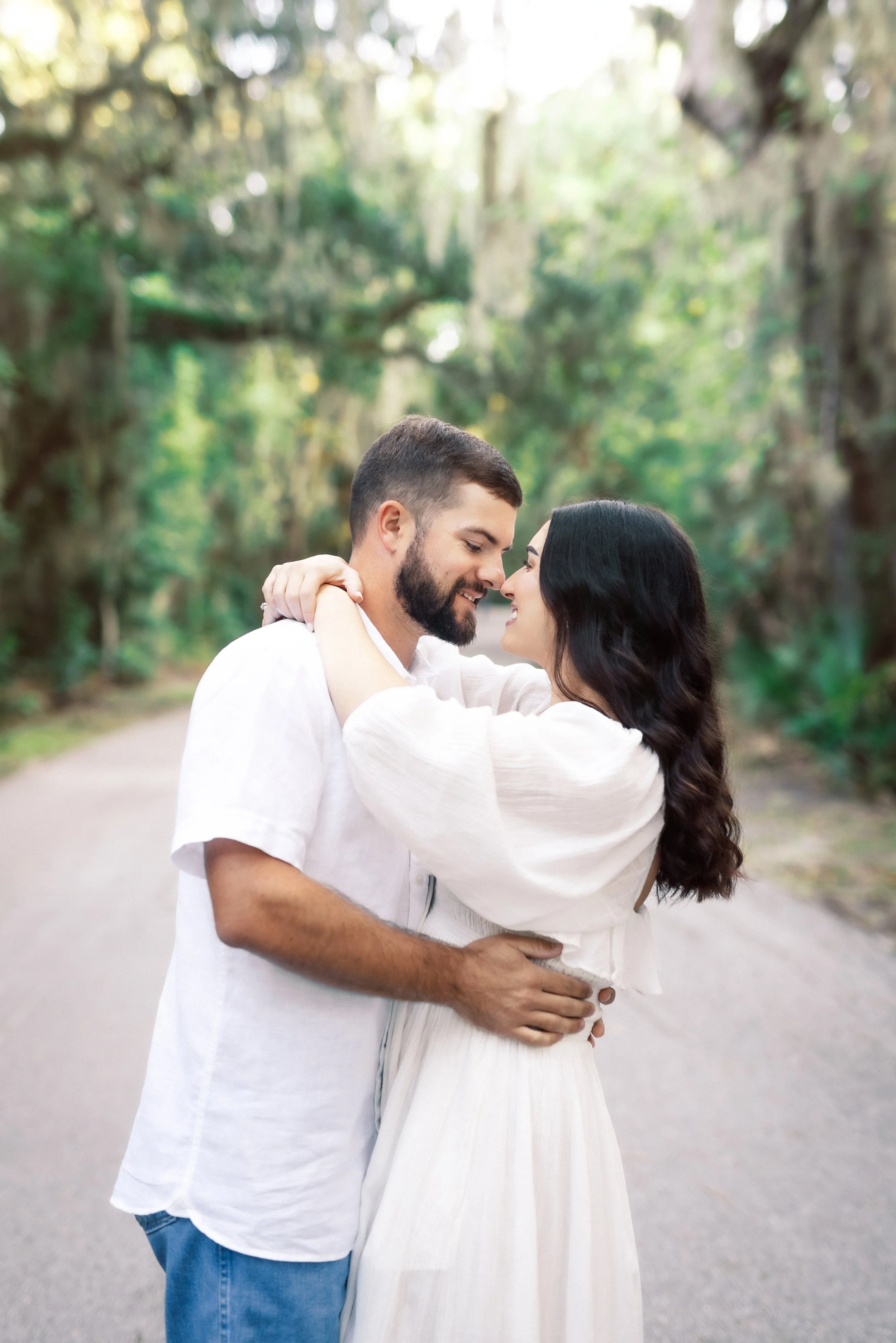A couple embracing outdoors on a path, with trees and greenery in the background, smiling and touching foreheads.