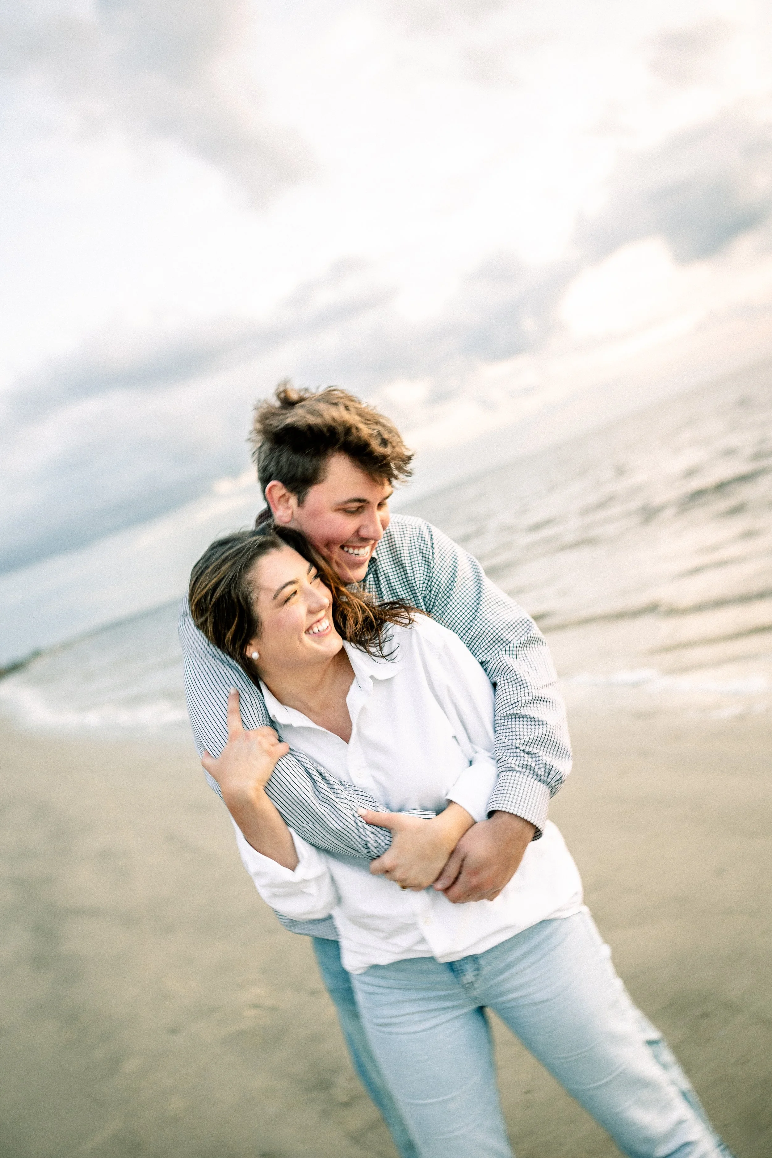 Two people, a man and a woman, smiling and hugging on a beach with cloudy skies in the background.