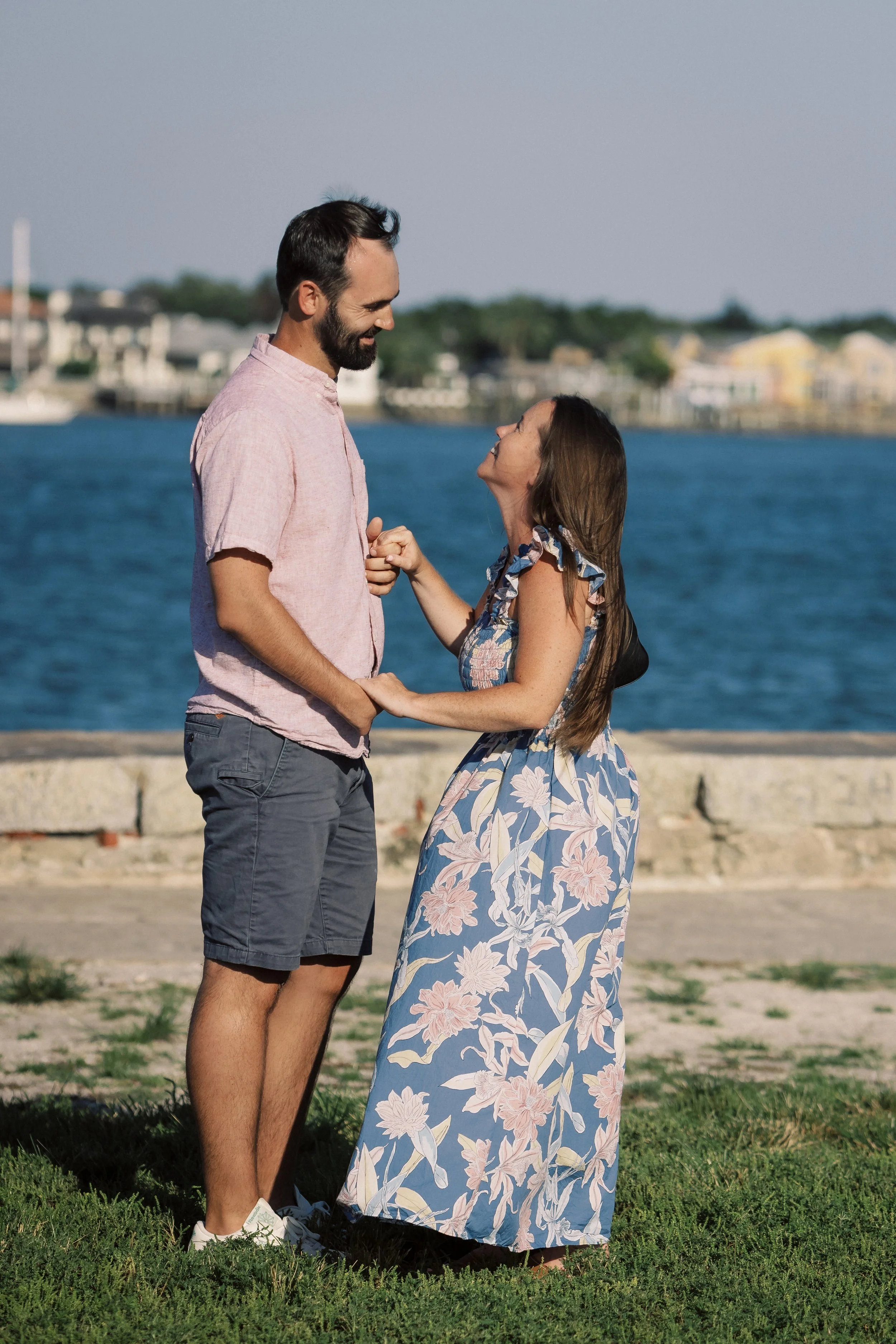 A couple standing close together near water, holding hands and smiling at each other with a cityscape in the background.