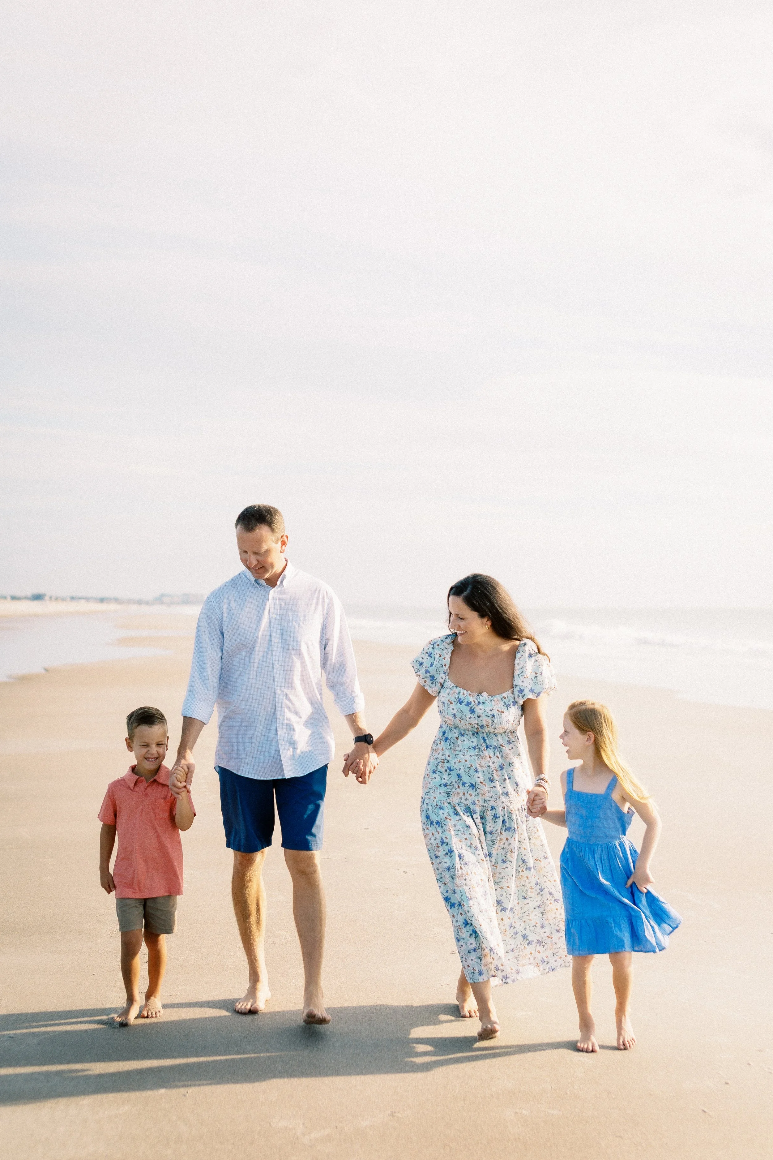 A family walking hand in hand on a sandy beach, smiling, with the ocean and sky in the background.Photographer Amelia Island, Jacksonville, St Augustine, St Simons Island, Savannah photographer