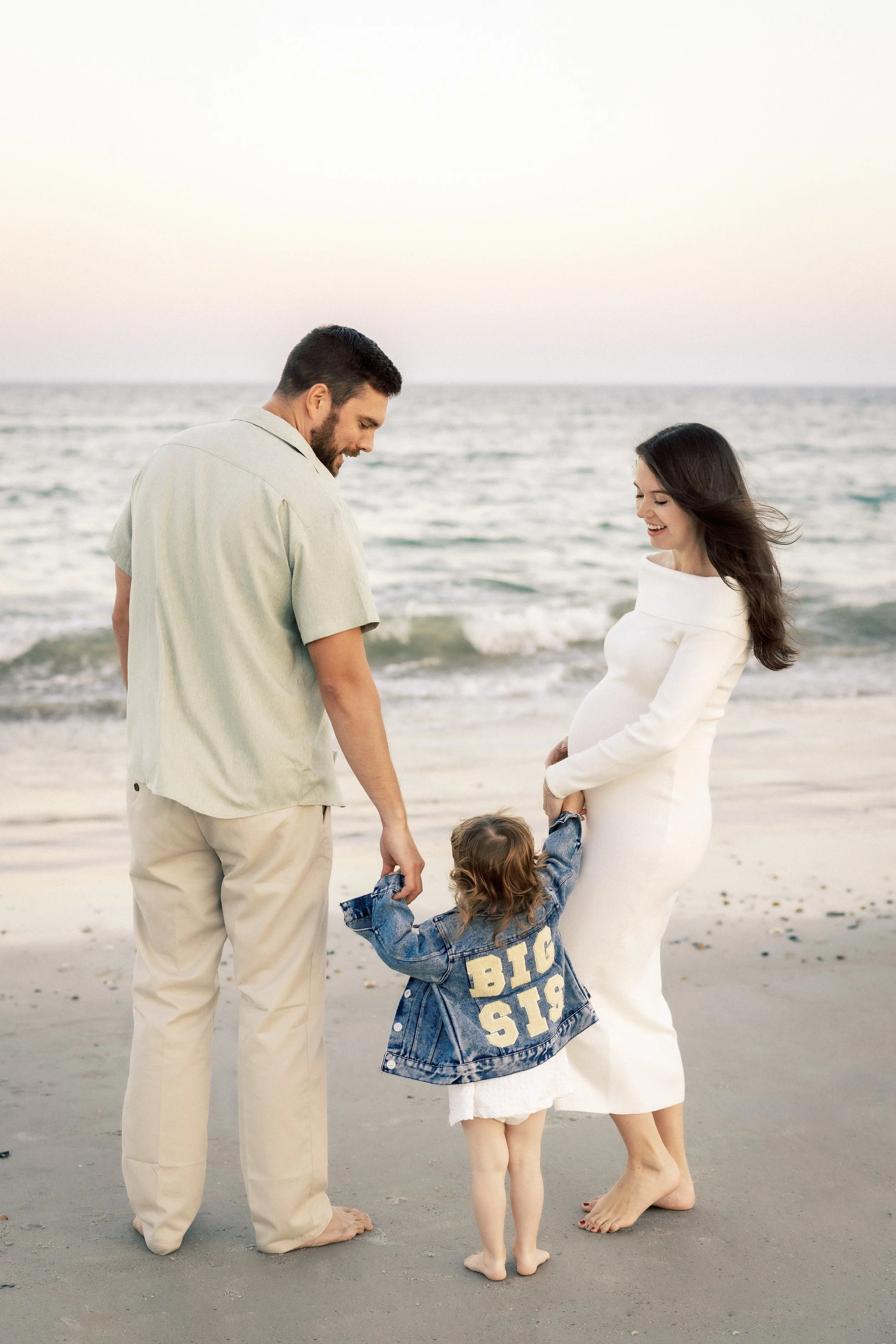 A family of four standing barefoot on the beach. The father and mother are holding hands with their two young children, a boy and a girl. The mother is pregnant and smiling, the father is looking down at the children, and the children are playing tog