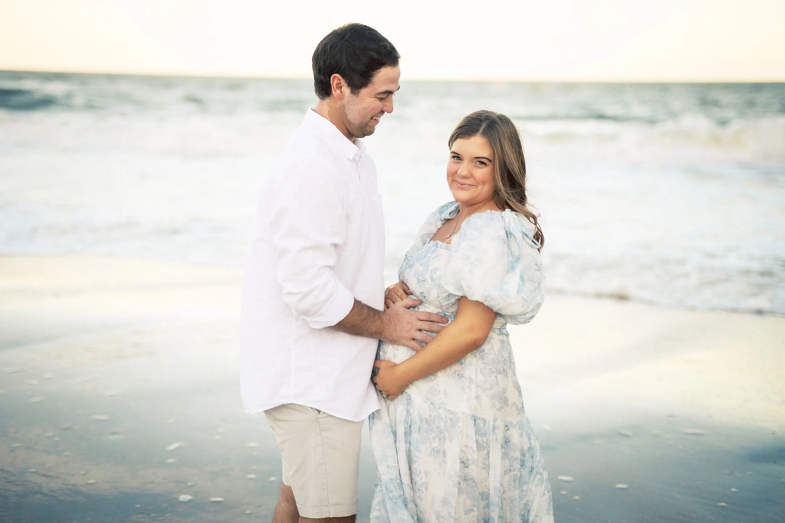 A couple standing on the beach, holding hands and smiling, with the ocean in the background.