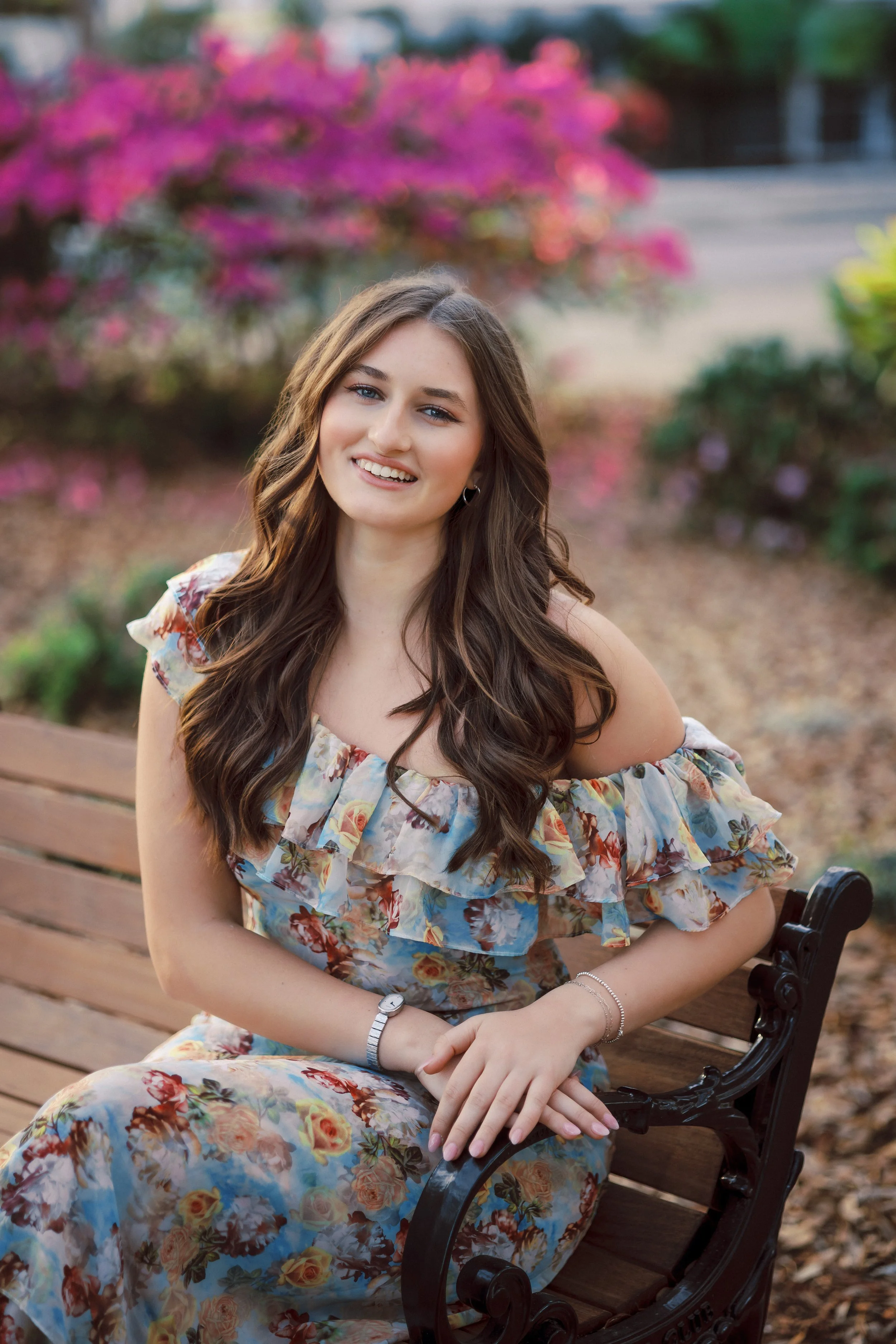 A young woman with long, wavy brown hair, wearing a floral dress, sitting on a wooden bench in a park with pink flowers and greenery in the background, smiling at the camera.