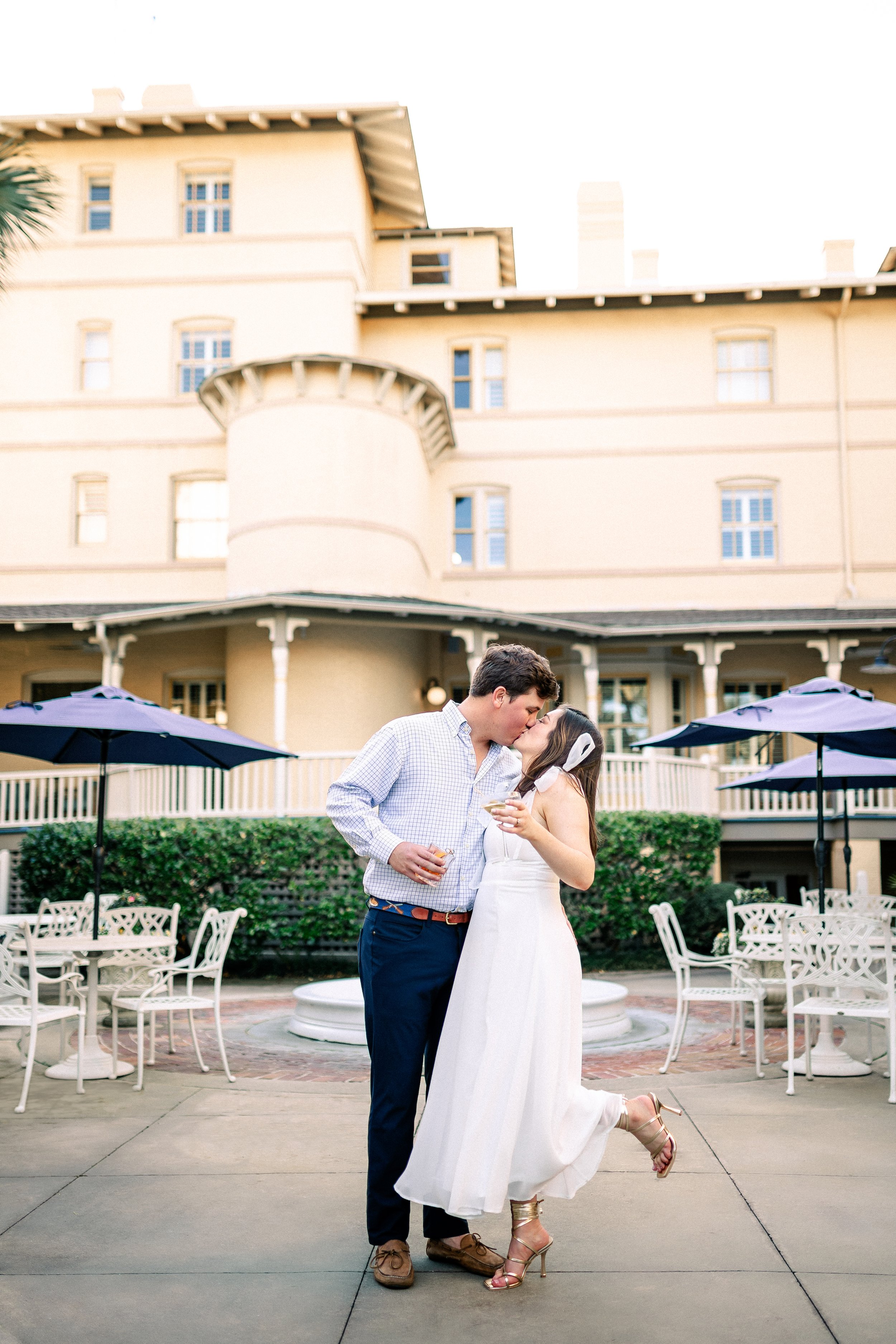 A bride and groom sharing a kiss at an outdoor wedding reception with white chairs, umbrellas, and a large historic building in the background.