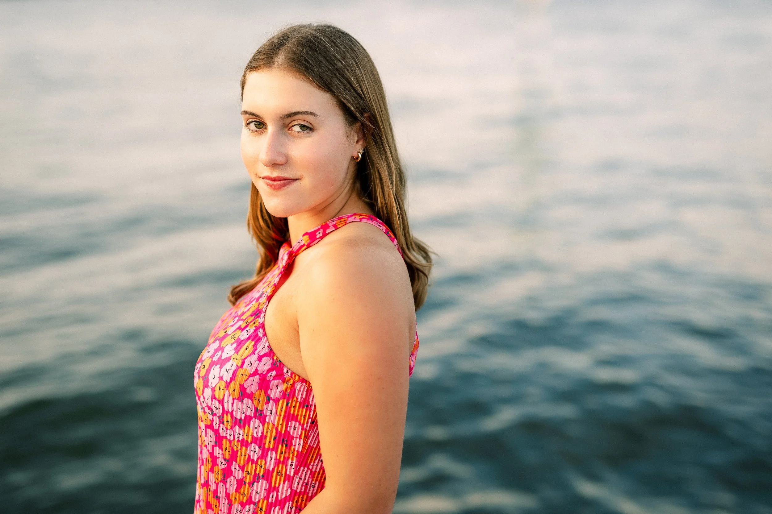 A young woman with light skin and brown hair looking over her shoulder at the camera, standing by a body of water with a calm, reflective surface, wearing a colorful, patterned sleeveless top.