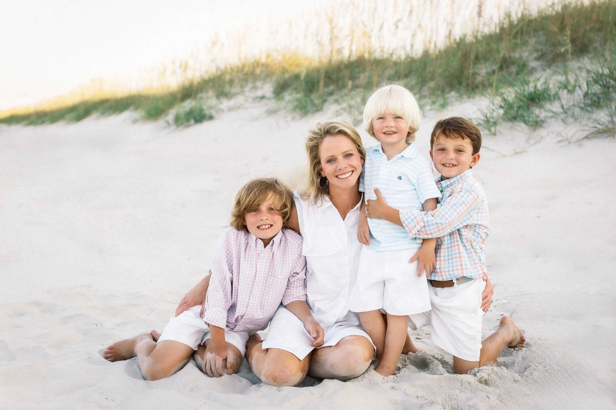 A woman with three young boys sits on the sand at the beach, smiling, with grass and sand dunes in the background. Photographer Amelia Island, Jacksonville, St Augustine, St Simons Island, Savannah photographer