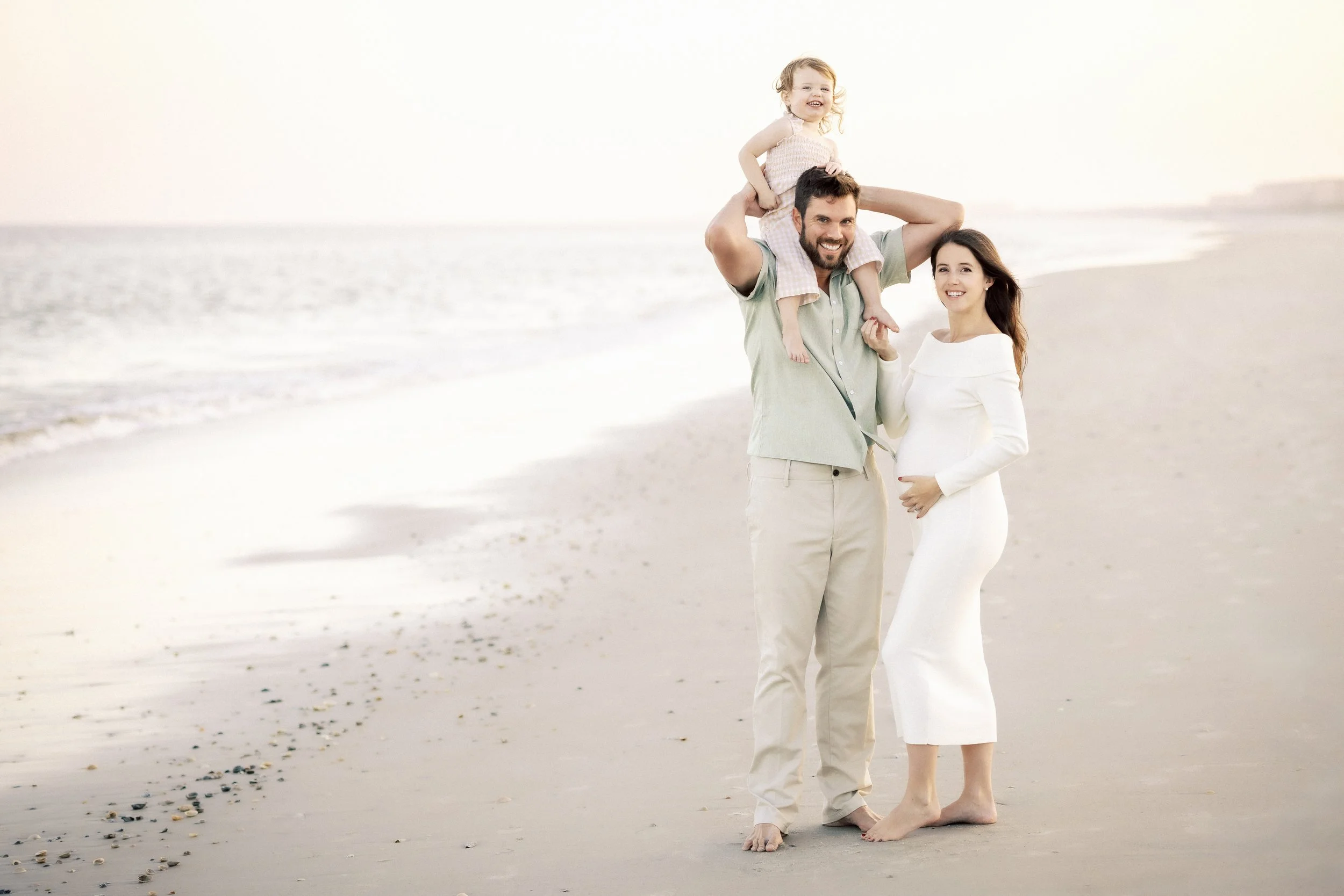 A happy family of three at the beach with a man carrying a small girl on his shoulders and a woman standing beside them, all smiling.Photographer Amelia Island, Jacksonville, St Augustine, St Simons Island, Savannah photographer