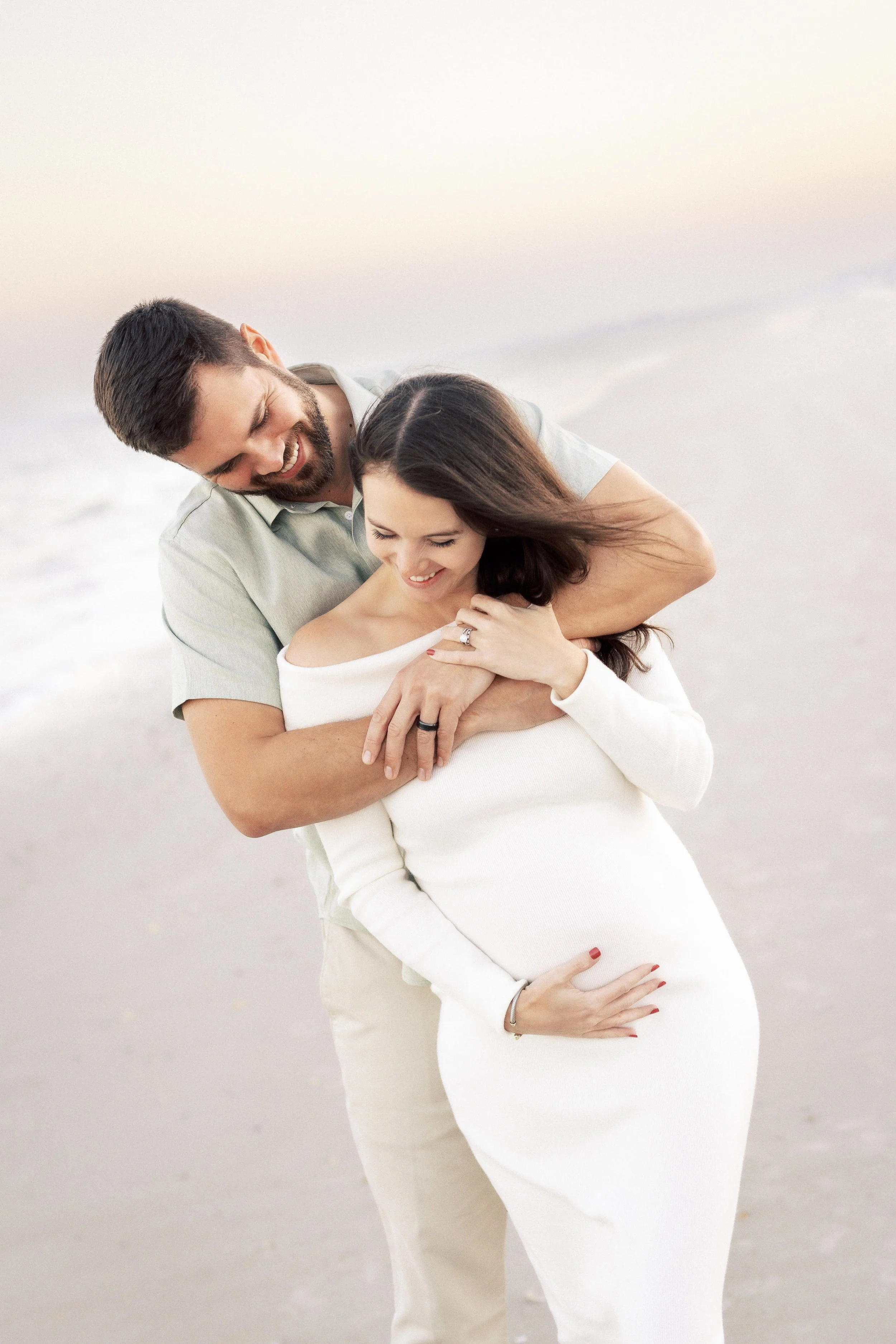 A man and a pregnant woman smiling and embracing on a beach during sunset.