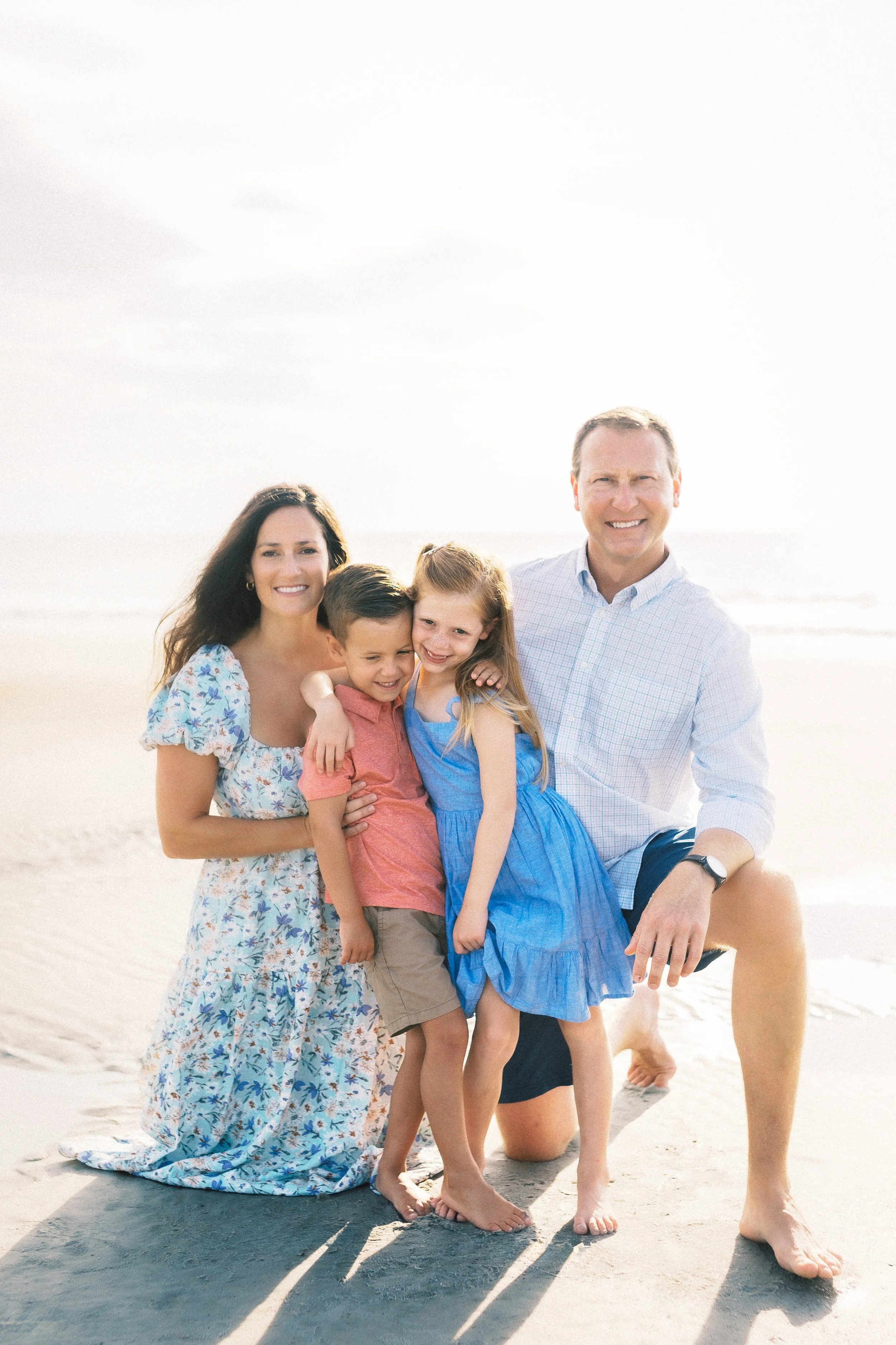 A family of four, two adults and two children, standing on a beach with sand and water in the background. They are smiling and embracing each other.