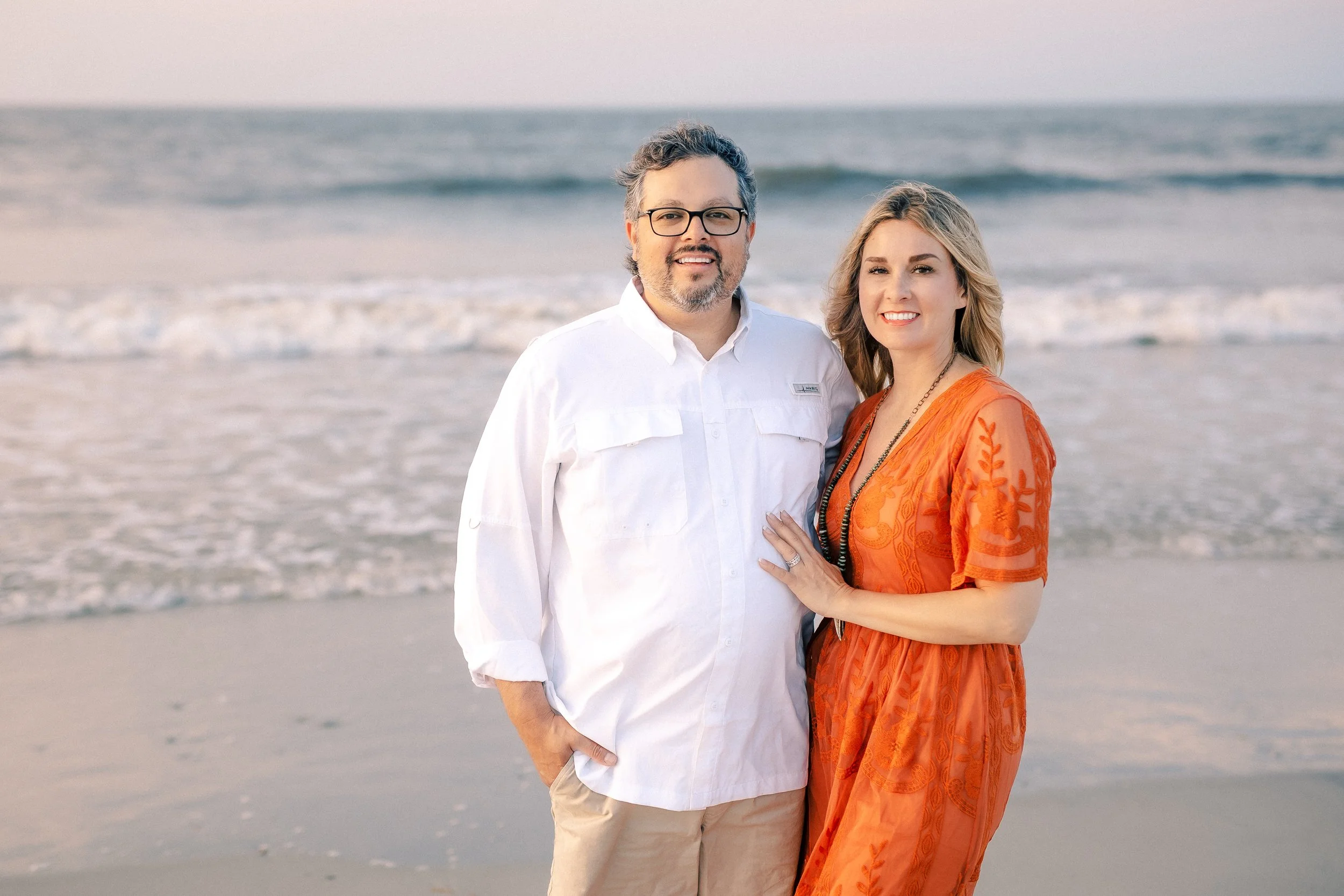 A man and woman standing on a beach with the ocean in the background. The man is wearing glasses, a white button-up shirt, and tan shorts. The woman is wearing an orange dress with embroidery and has her hand on the man's arm.