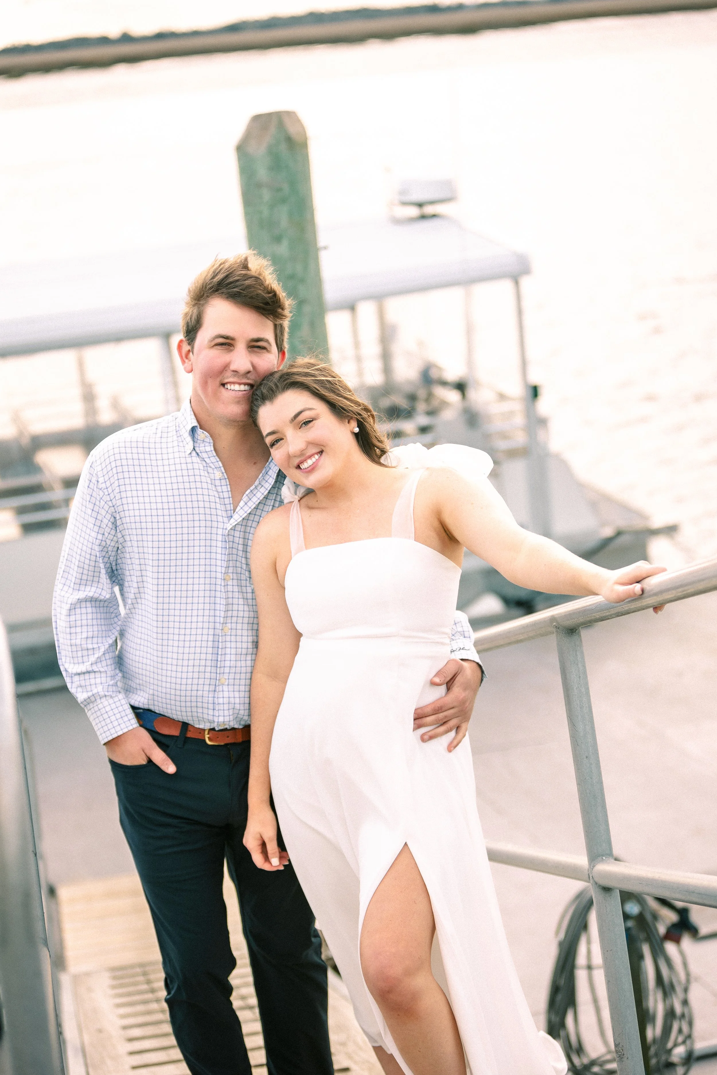 A happy couple standing outdoors near a body of water and a boat, smiling at the camera.