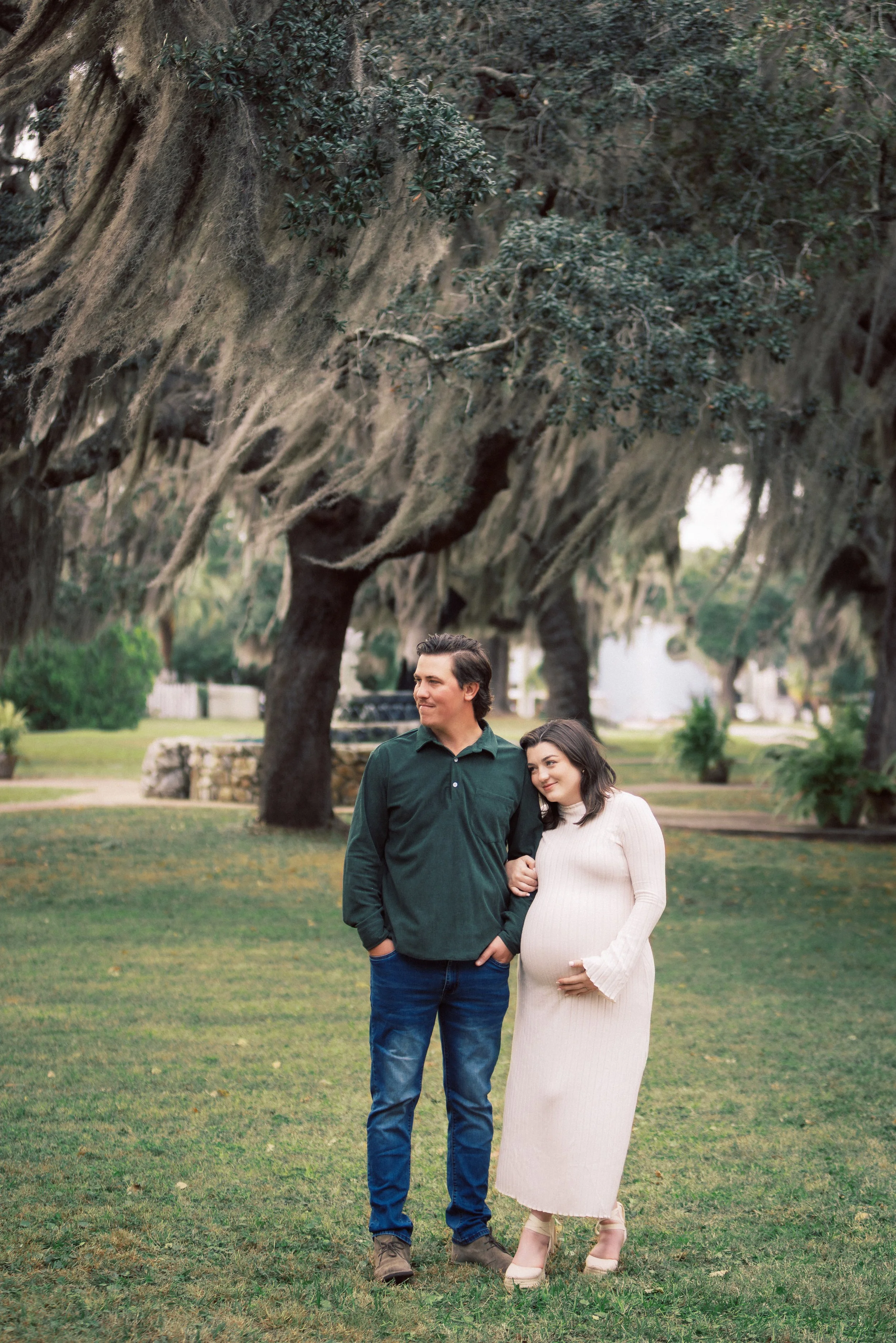 A pregnant woman with dark hair in a white long sleeve dress holding her belly, stands arm in arm with a man in a green button-up shirt and blue jeans. They are outside on a grassy area with large trees covered in Spanish moss in the background.