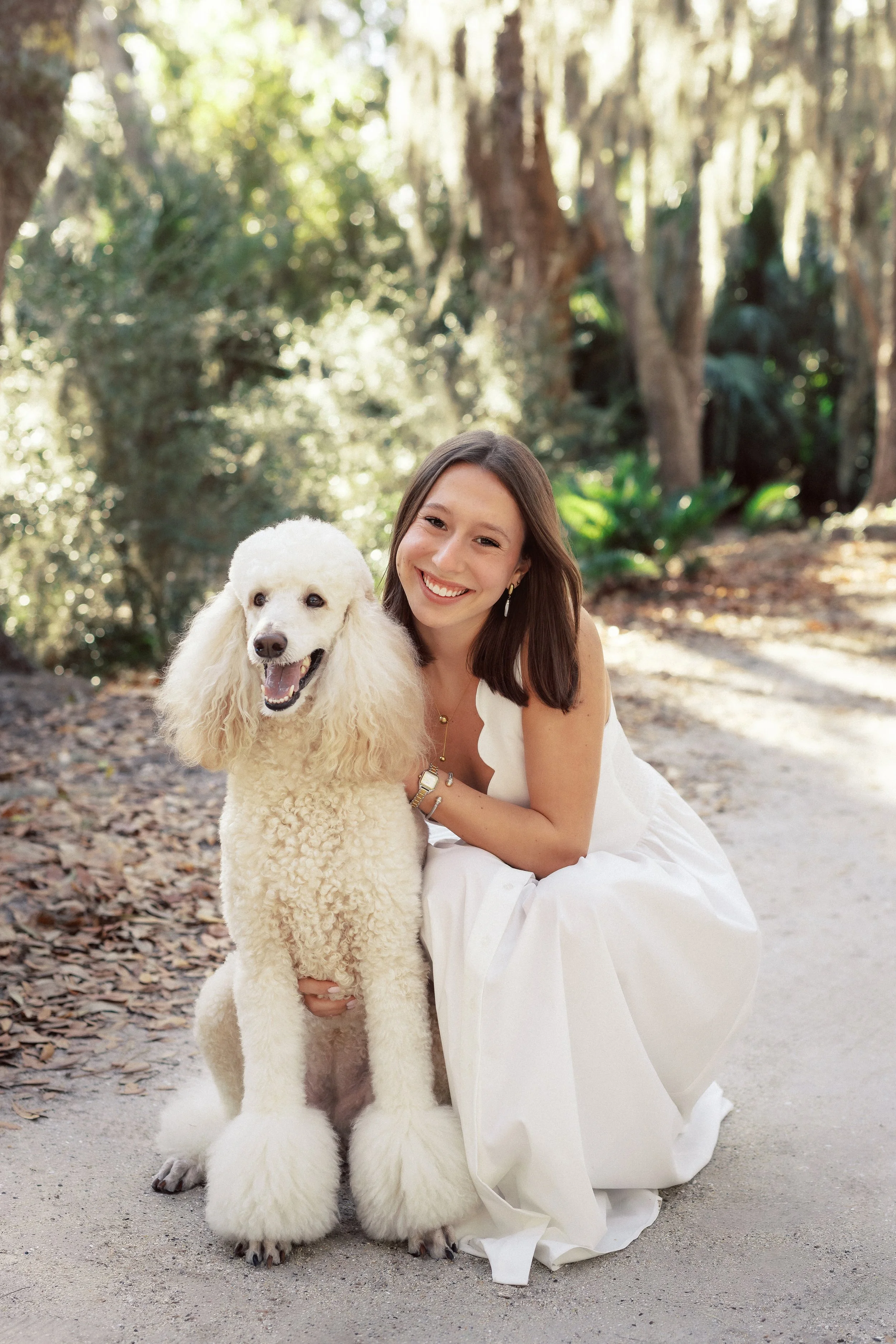 A woman in a white dress kneels beside a large, white poodle with grooming that resembles a lion, on a forest trail with trees and greenery in the background.