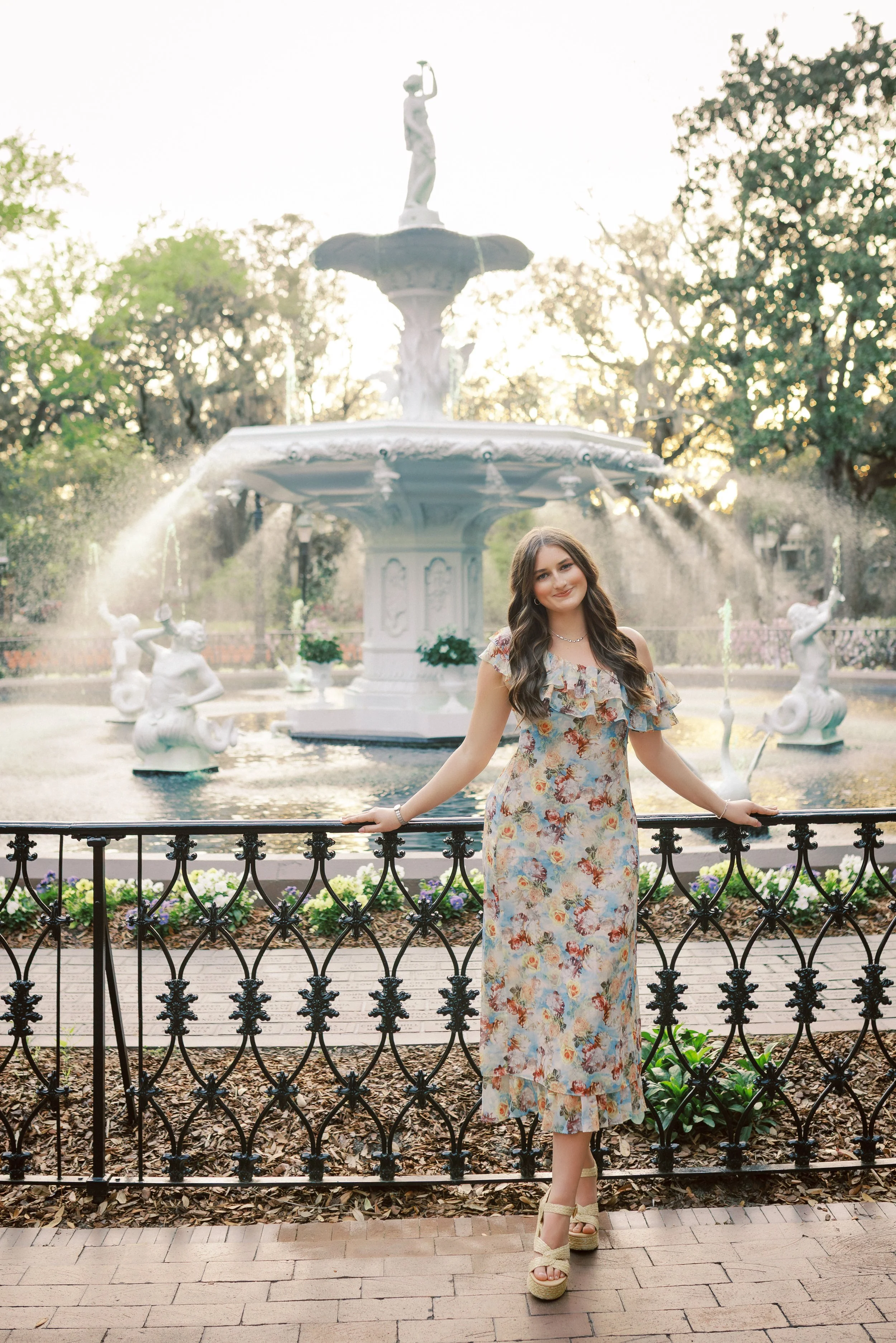 A young woman in a floral dress standing near a decorative black iron fence in front of a fountain with a statue on top, in a park with trees and sunlight.