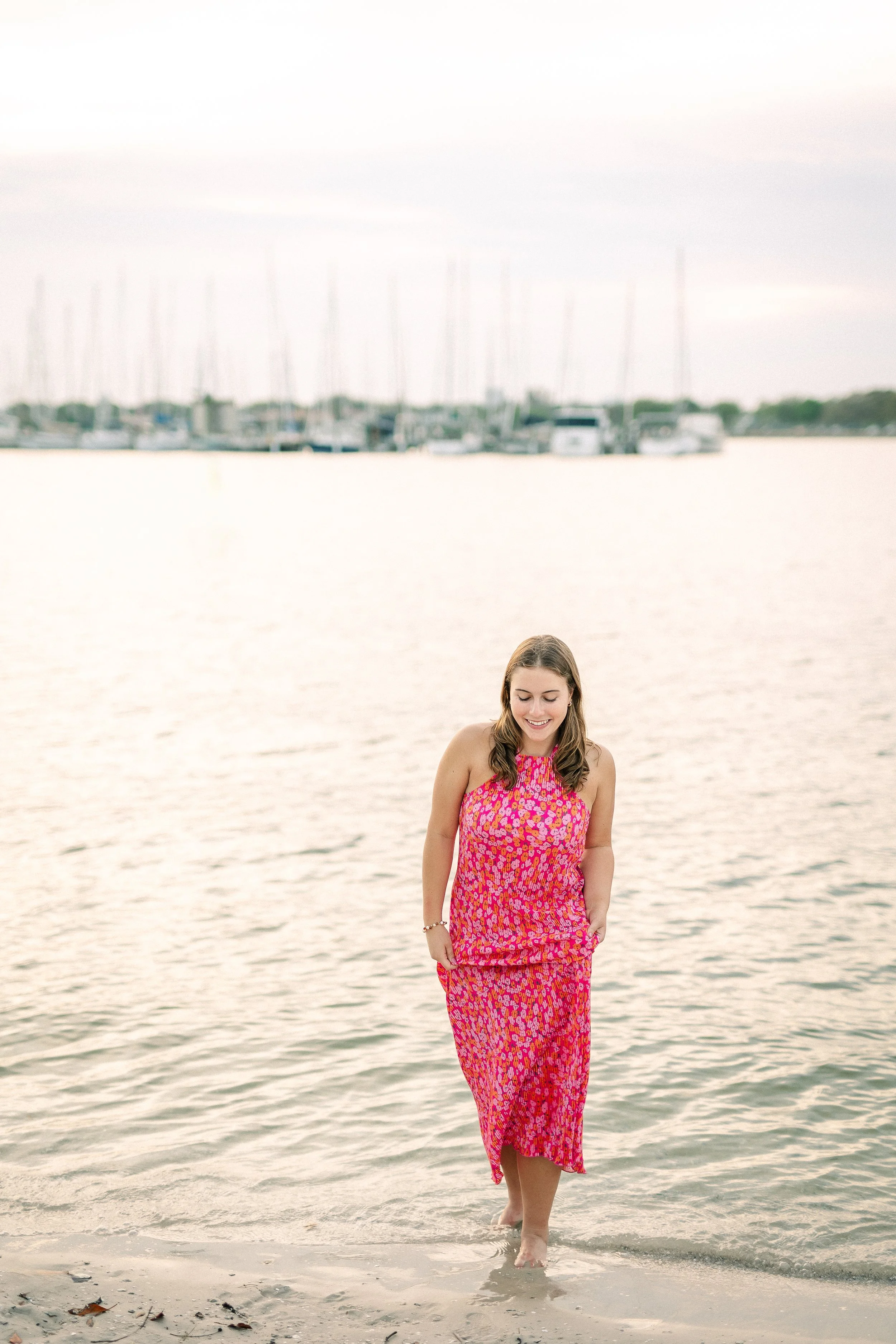 A woman in a pink and purple patterned dress walking barefoot along the shoreline of a beach, with water and a marina with boats in the background.