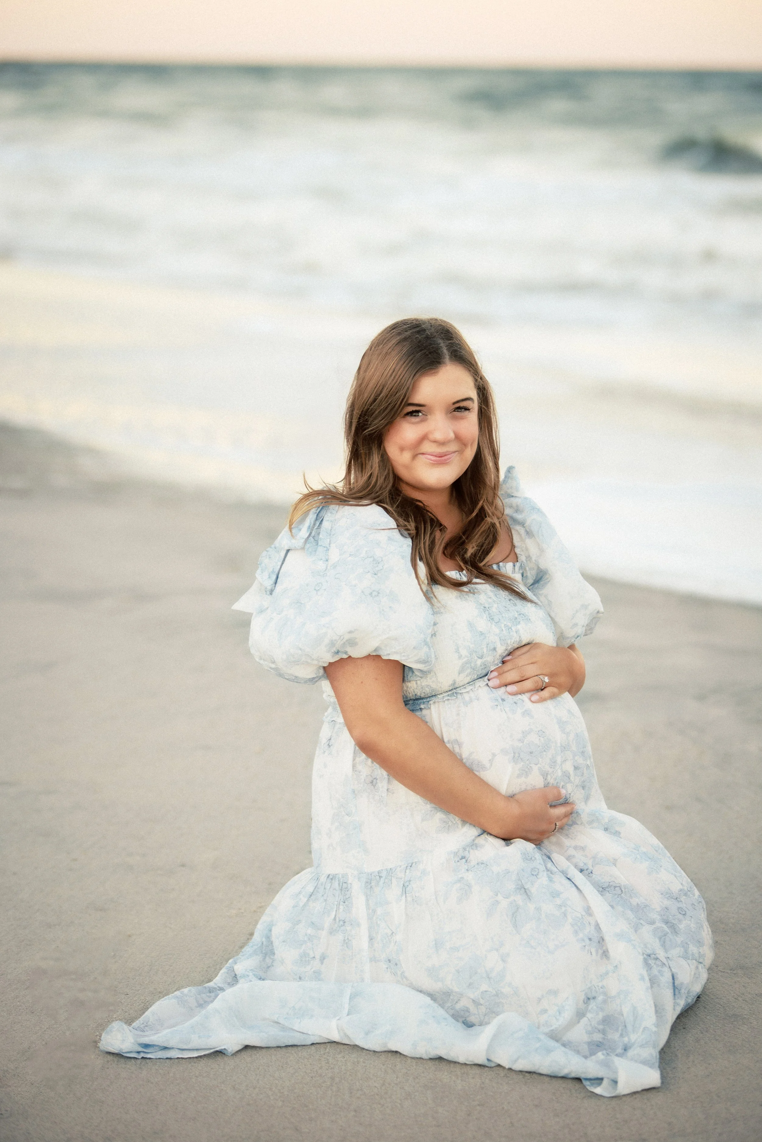 A pregnant woman with long brown hair, wearing a light blue patterned dress, sitting on the beach with the ocean in the background, smiling and looking at the camera.