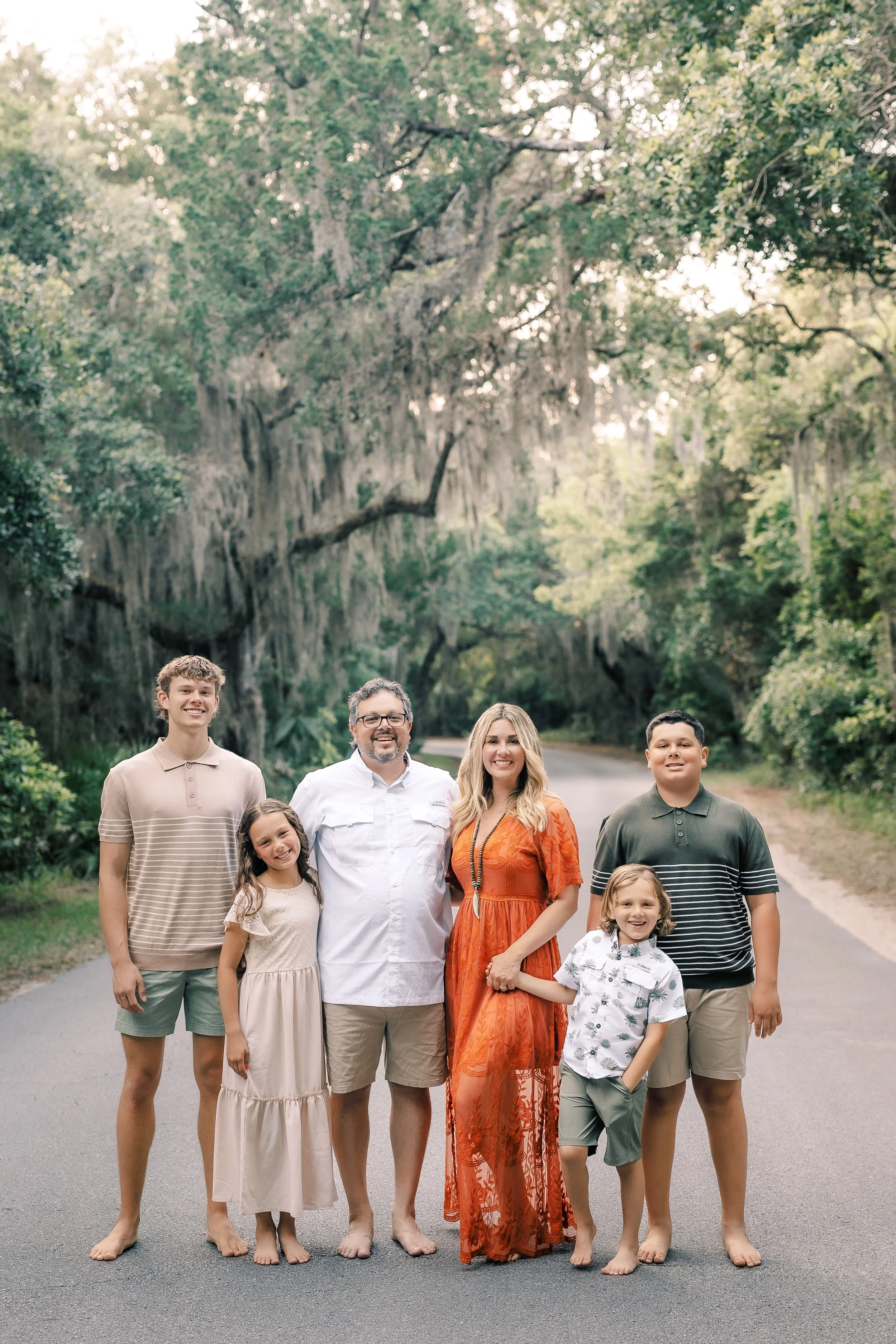 A large family standing together on a road in a wooded area with tall trees and Spanish moss overhead, smiling at the camera.