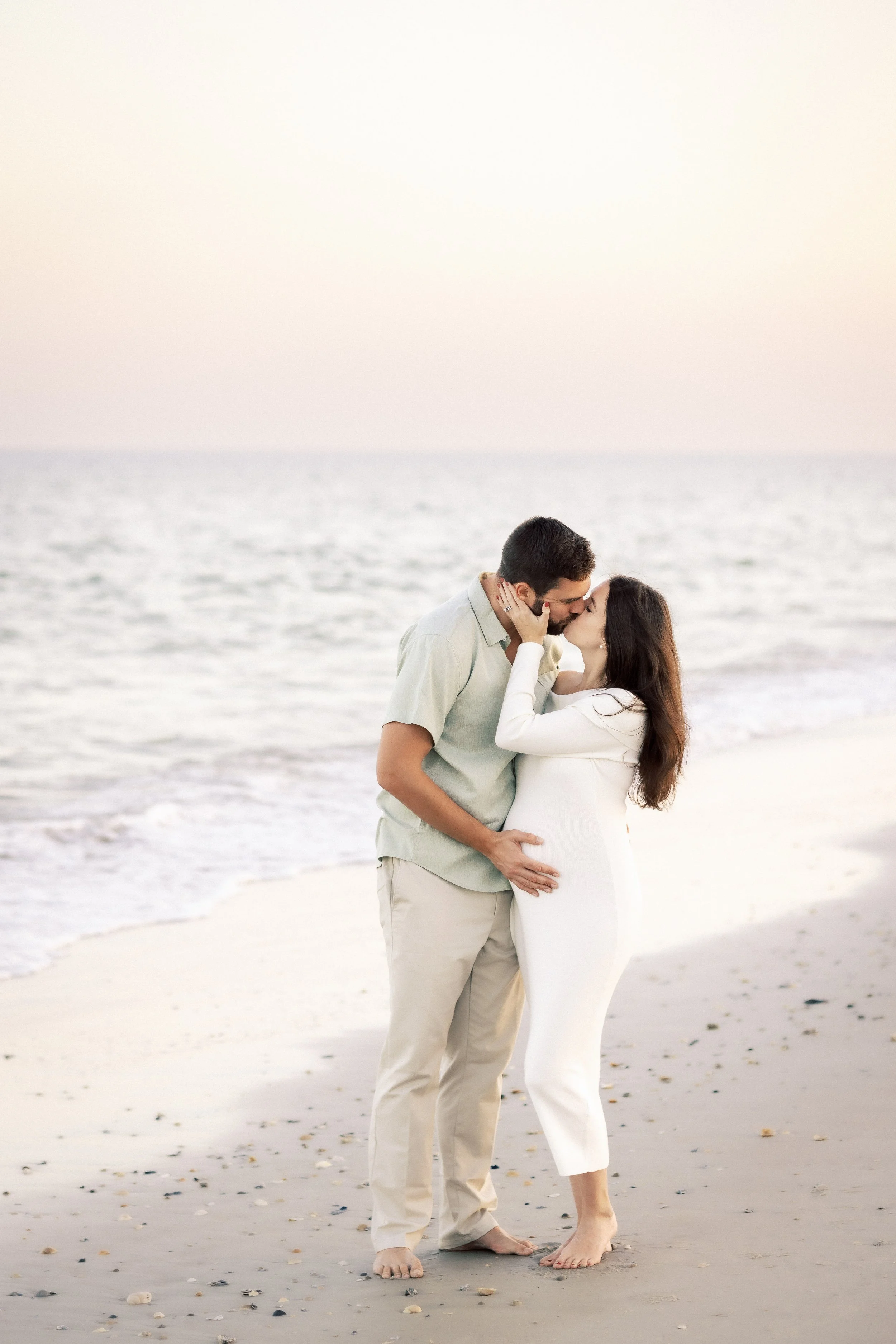 A couple is sharing a kiss on the beach at sunset, with ocean waves in the background and the woman holding her pregnant belly.Photographer Amelia Island, Jacksonville, St Augustine, St Simons Island, Savannah photographer