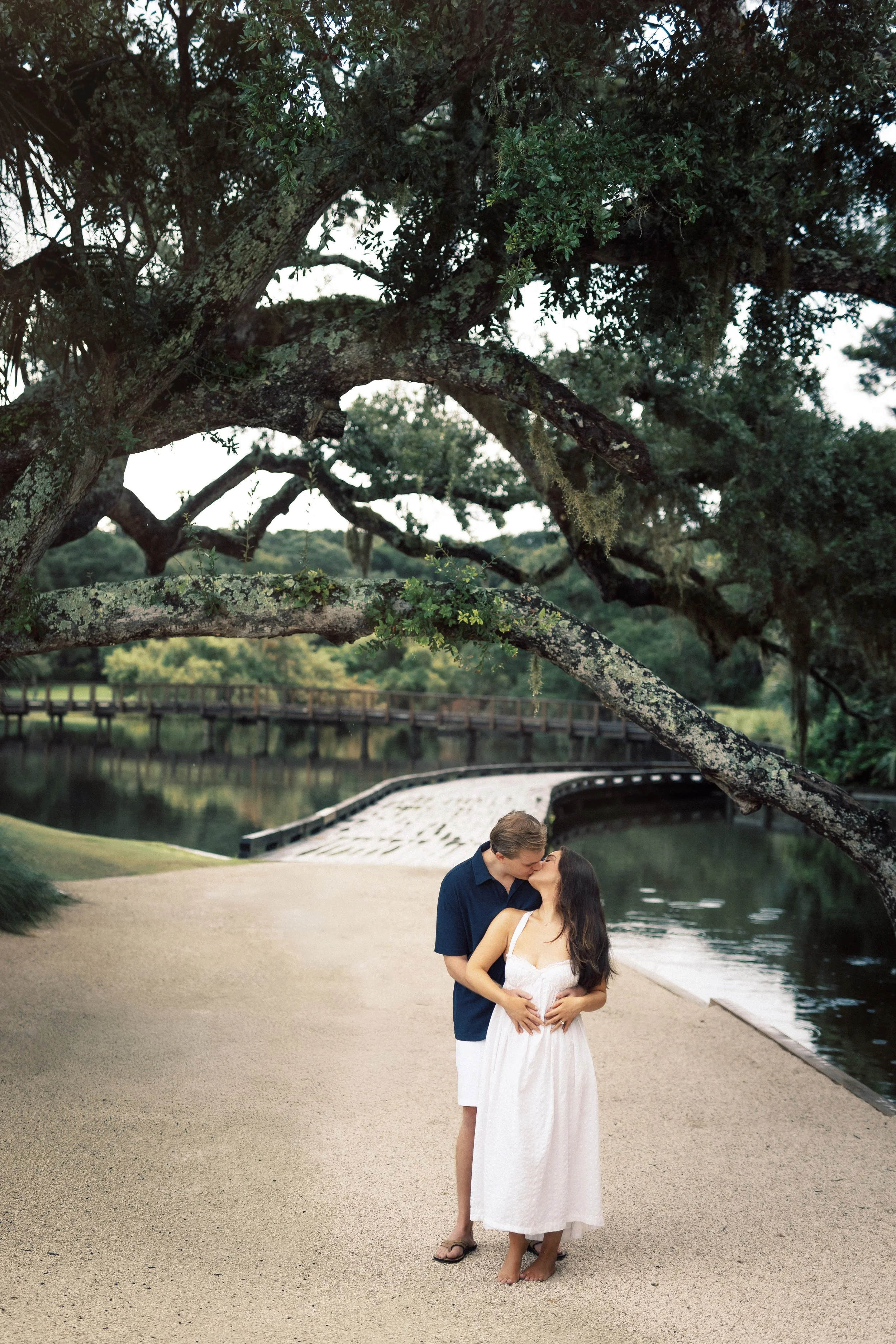 A romantic couple kissing on a riverbank, framed by sprawling tree branches with hanging moss, in a park with a footbridge and calm water in the background.
