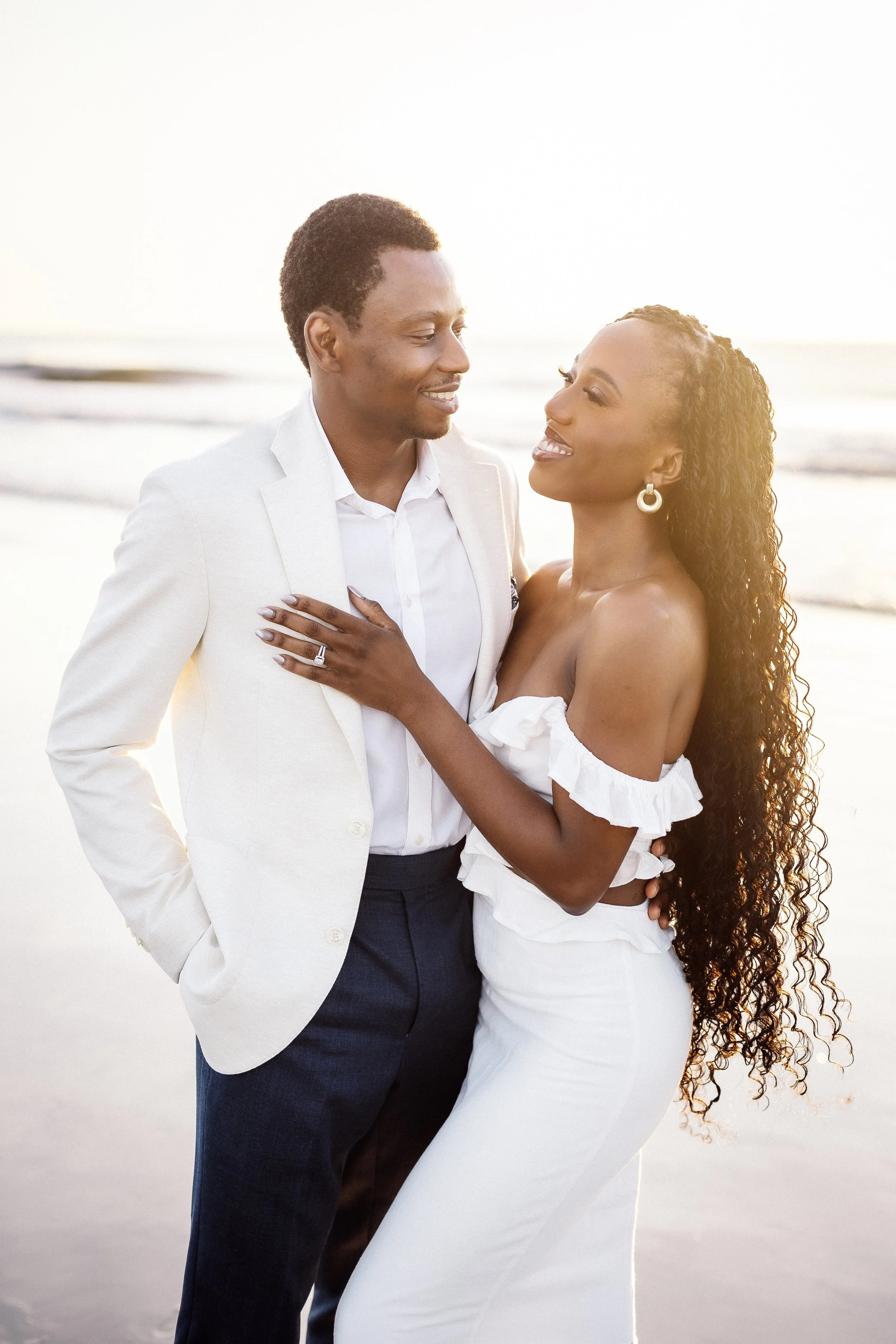A joyful interracial couple at the beach, smiling and embracing each other during sunset, dressed in formal white and navy attire.