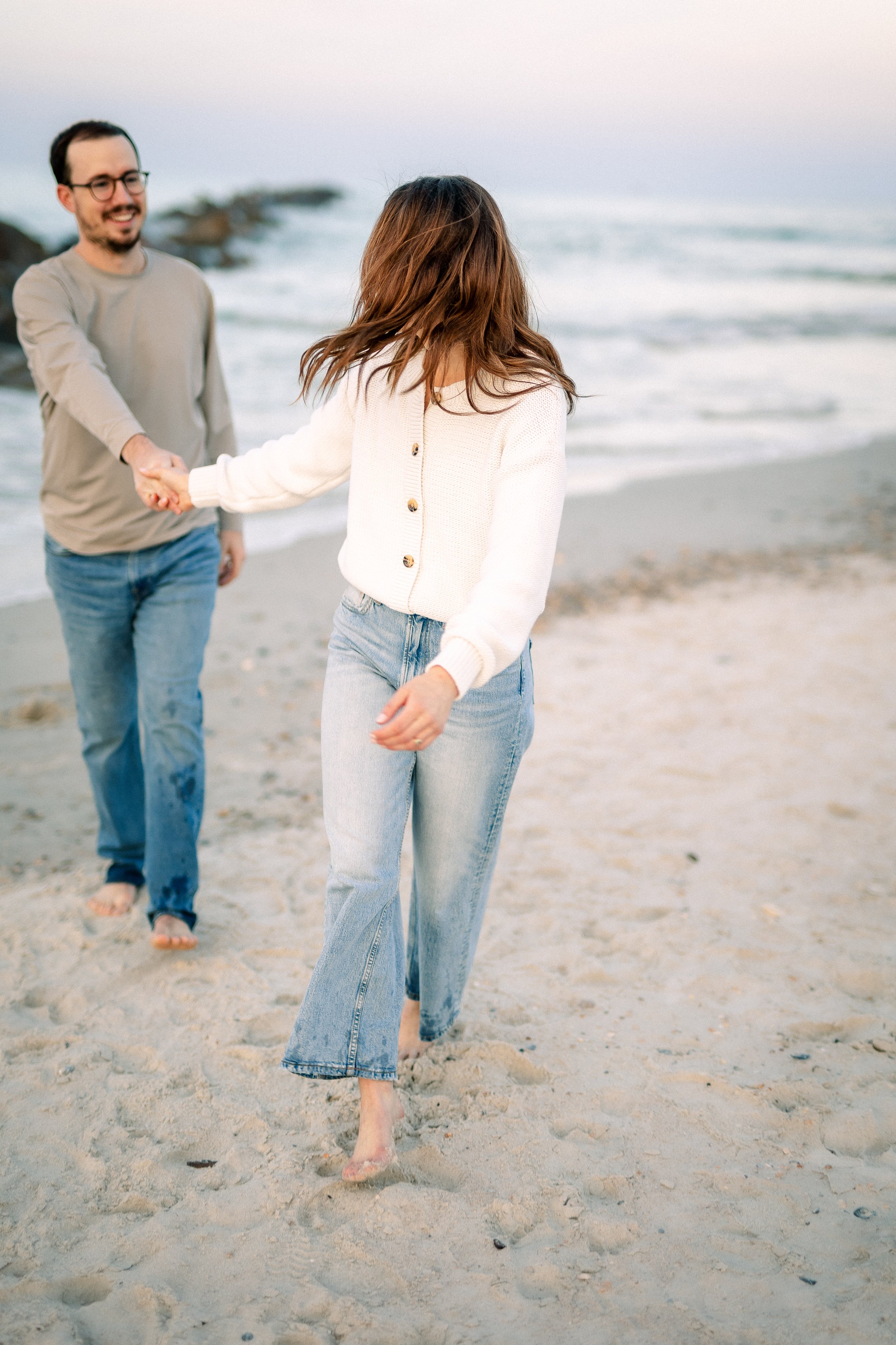 A woman and a man walk on a sandy beach, holding hands and smiling, with waves and rocks in the background.