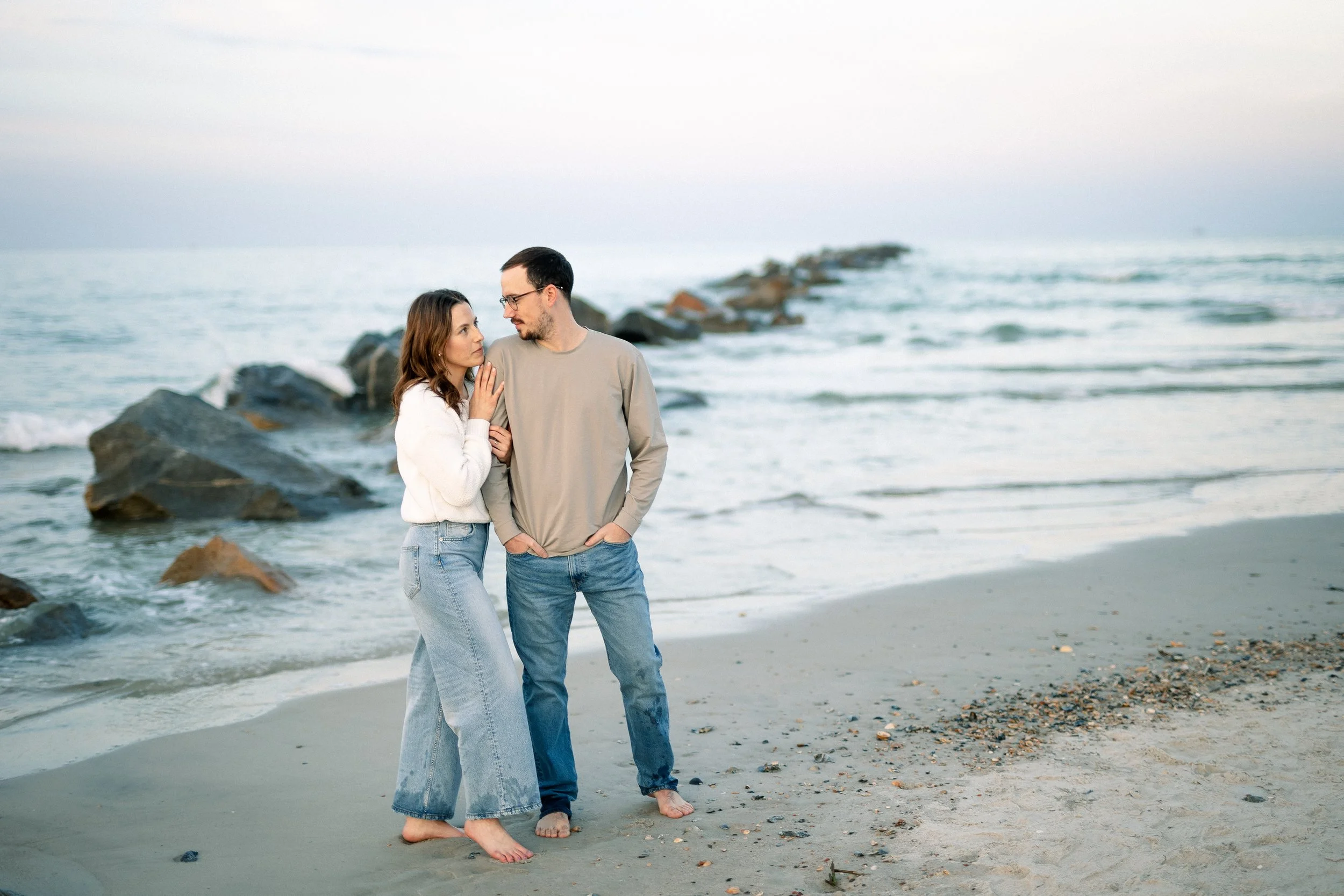 A couple walking barefoot on a sandy beach by the ocean, with rocks in the background, during daytime.