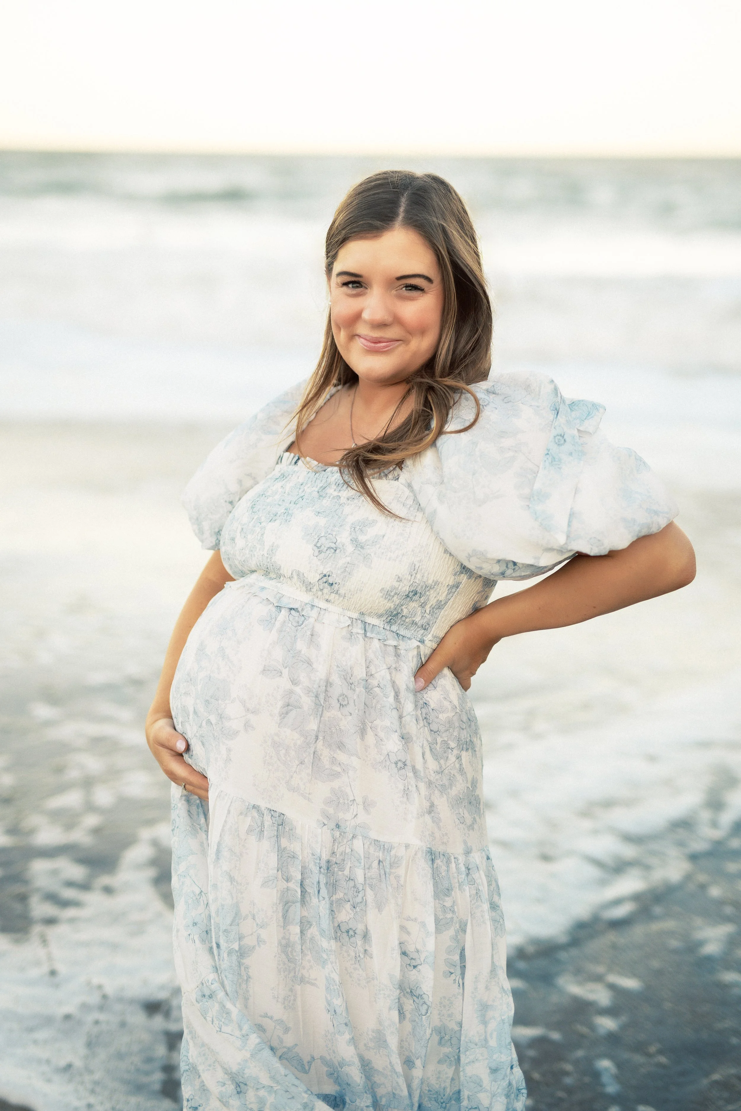 Pregnant woman standing on the beach, smiling, wearing a light-colored floral dress, with ocean waves and a cloudy sky in the background.