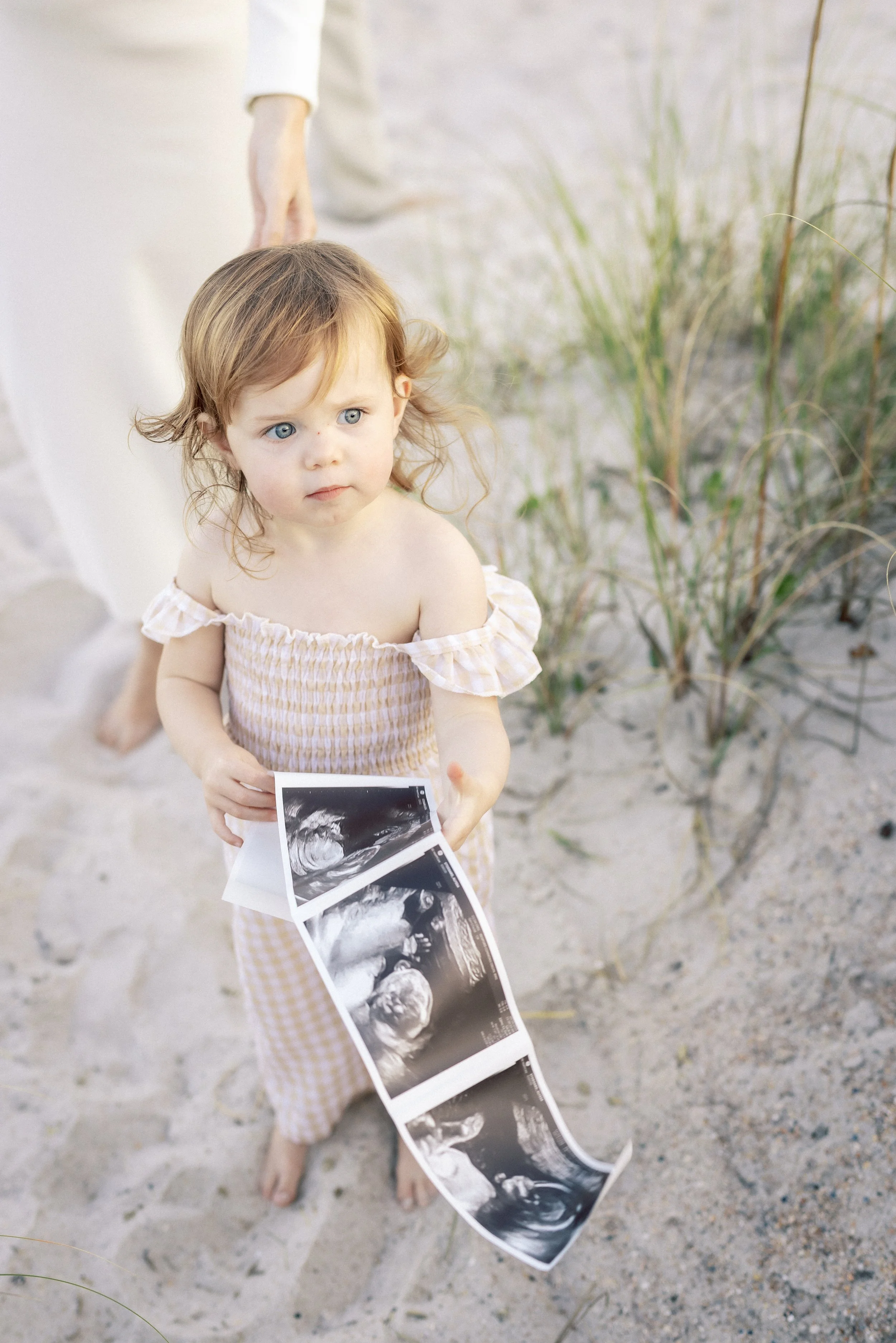 A young girl standing on sandy ground, holding ultrasound images, with taller grass in the background, and an adult hand partially visible nearby.Photographer Amelia Island, Jacksonville, St Augustine, St Simons Island, Savannah photographer