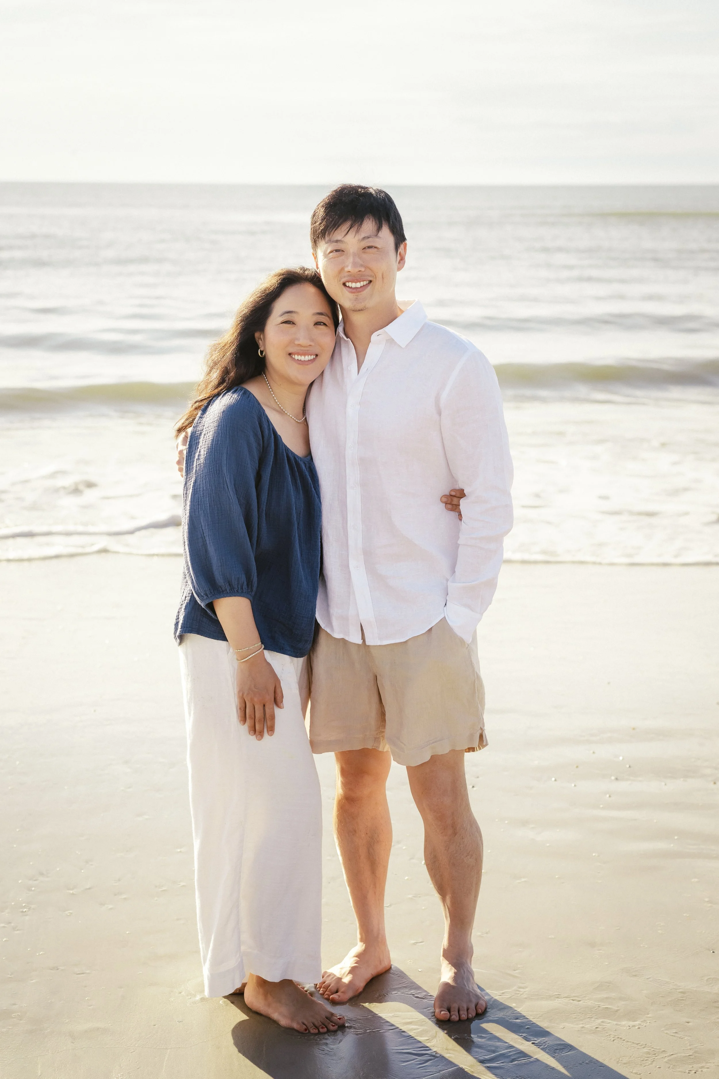 A smiling Asian couple standing barefoot on the beach, with waves in the background. The woman has long dark hair and is wearing a blue top and white skirt. Photographer Amelia Island, Jacksonville, St Augustine, St Simons Island, Savannah photograph