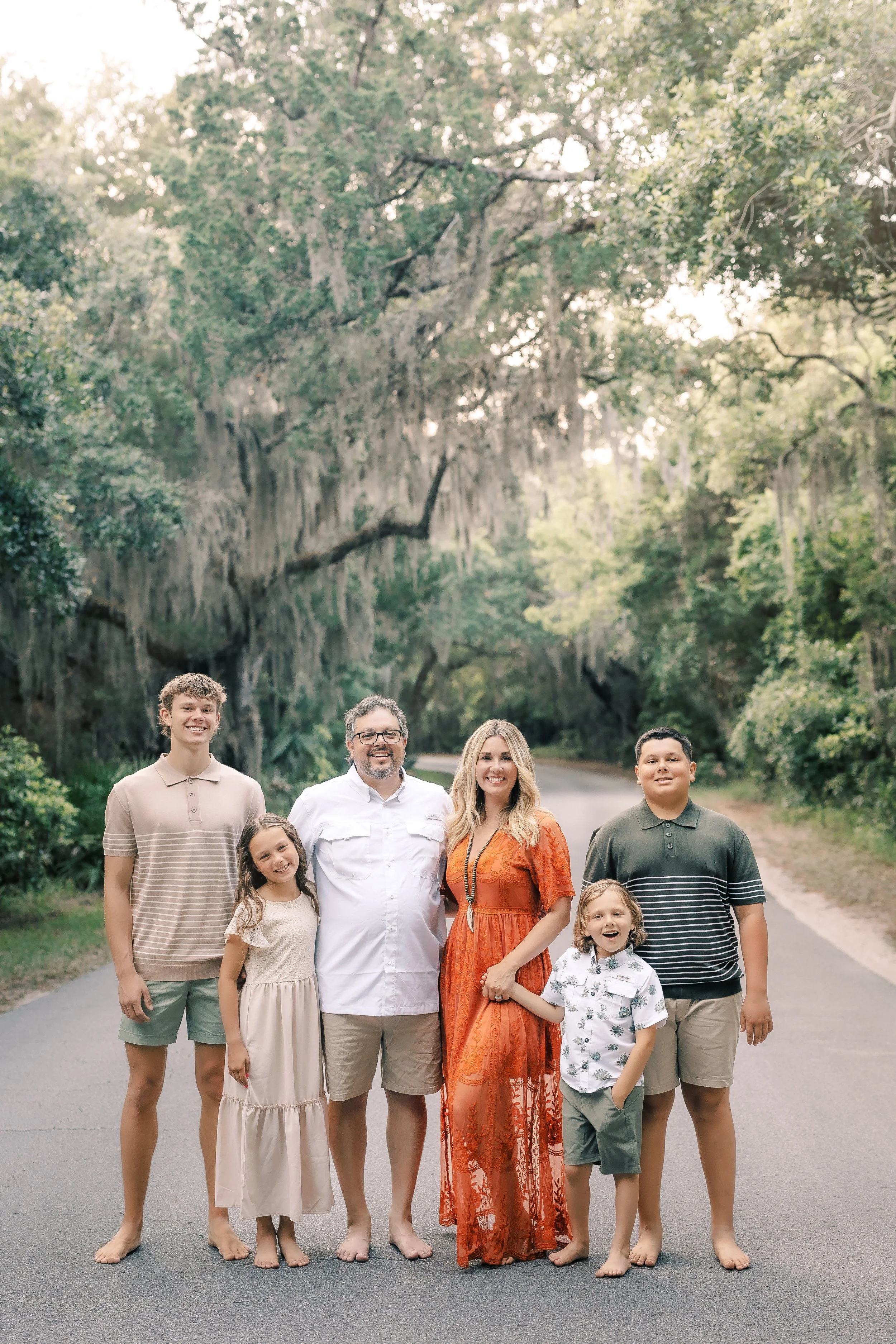 A group of seven people standing on a road surrounded by greenery, smiling at the camera. The group includes men, women, and children of various ages.