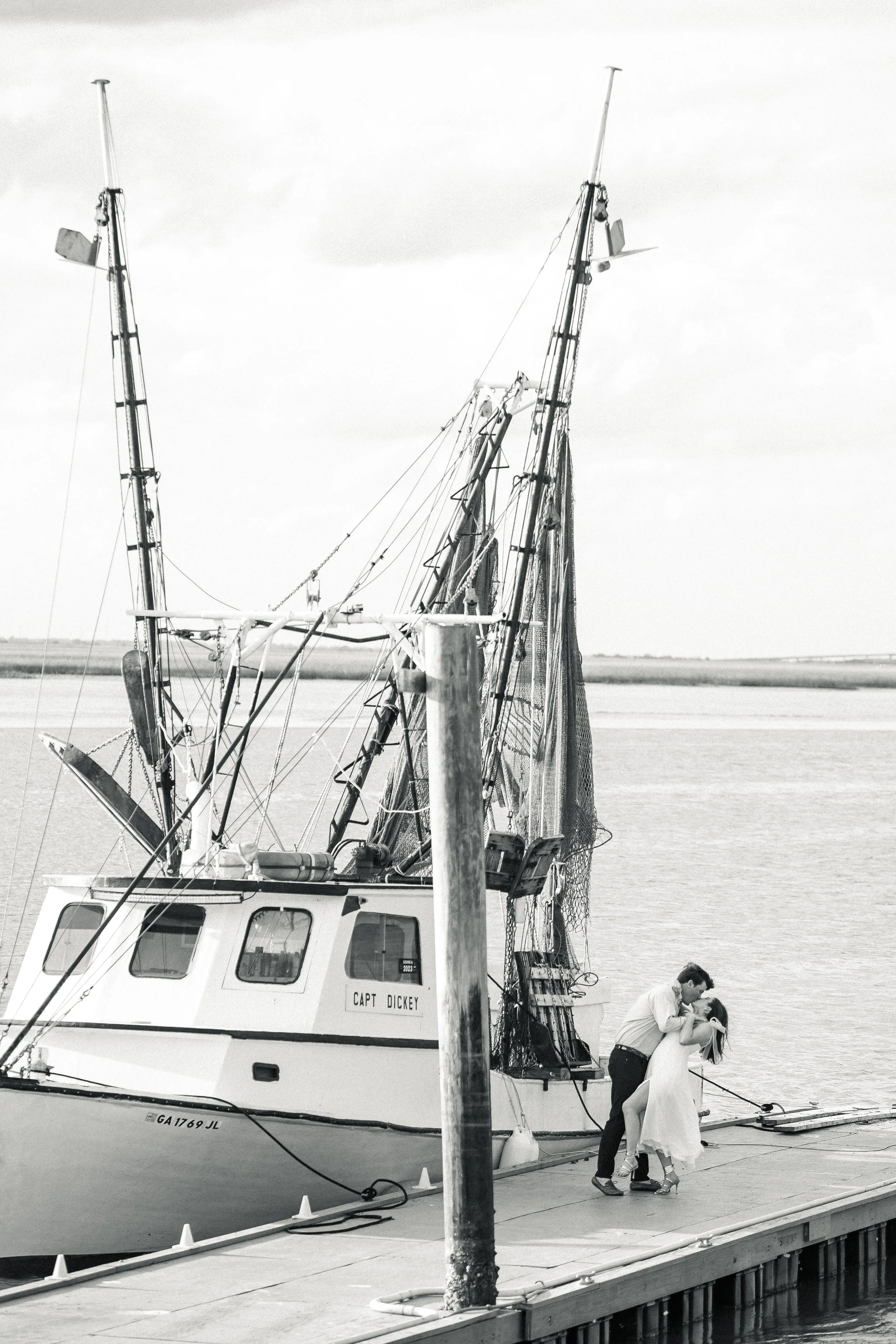 A man and woman dancing on a dock next to a boat, with the man dipping the woman. The boat has the name 'Capt. Dickey' on it and is docked by a body of water.