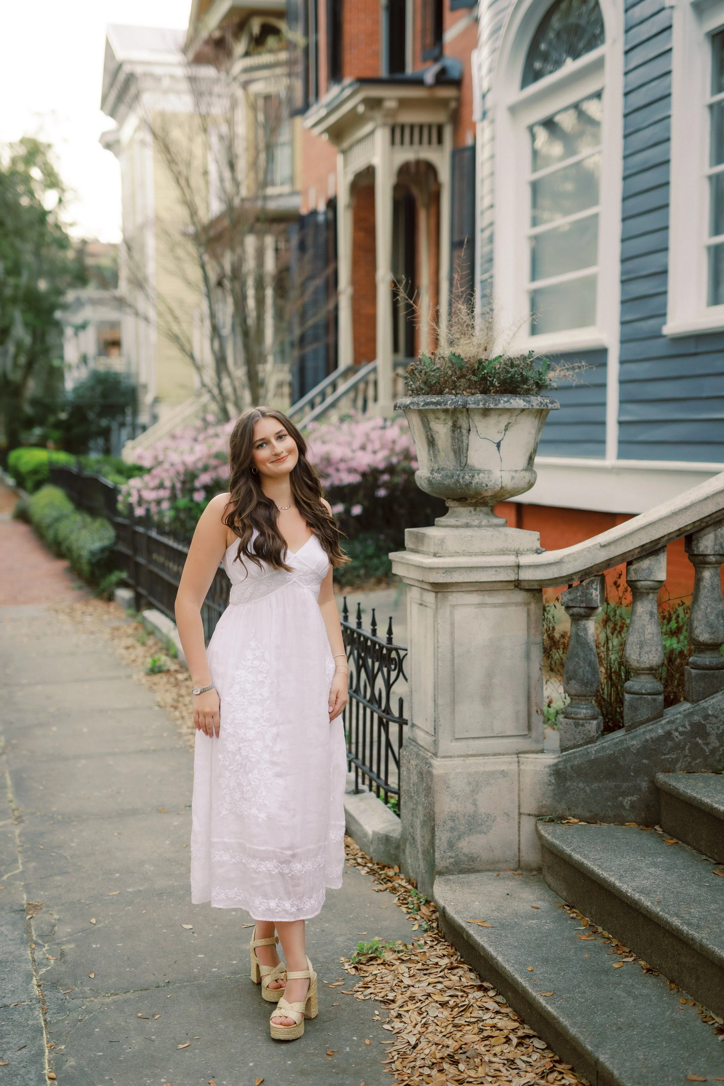 Young woman in a white dress standing on a sidewalk next to a stone staircase with a historic-style wrought iron fence and potted plant, in front of colorful row houses with bushes and trees in the background.
