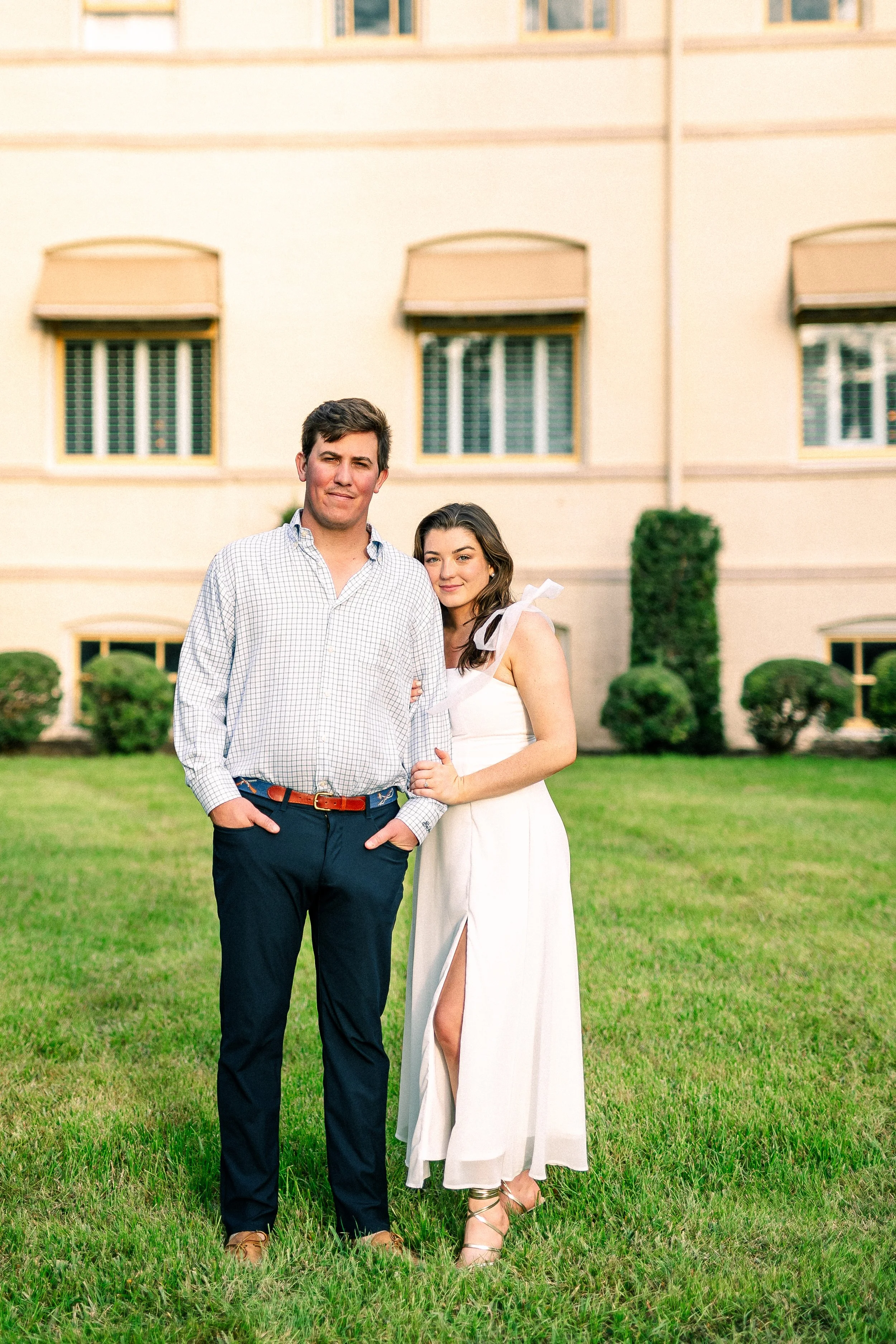 A young man and woman standing on a lawn in front of a beige building with multiple windows. The man wears a checkered shirt and dark pants, while the woman wears a white dress with high slits and strappy heels. They are smiling, with the woman's arm