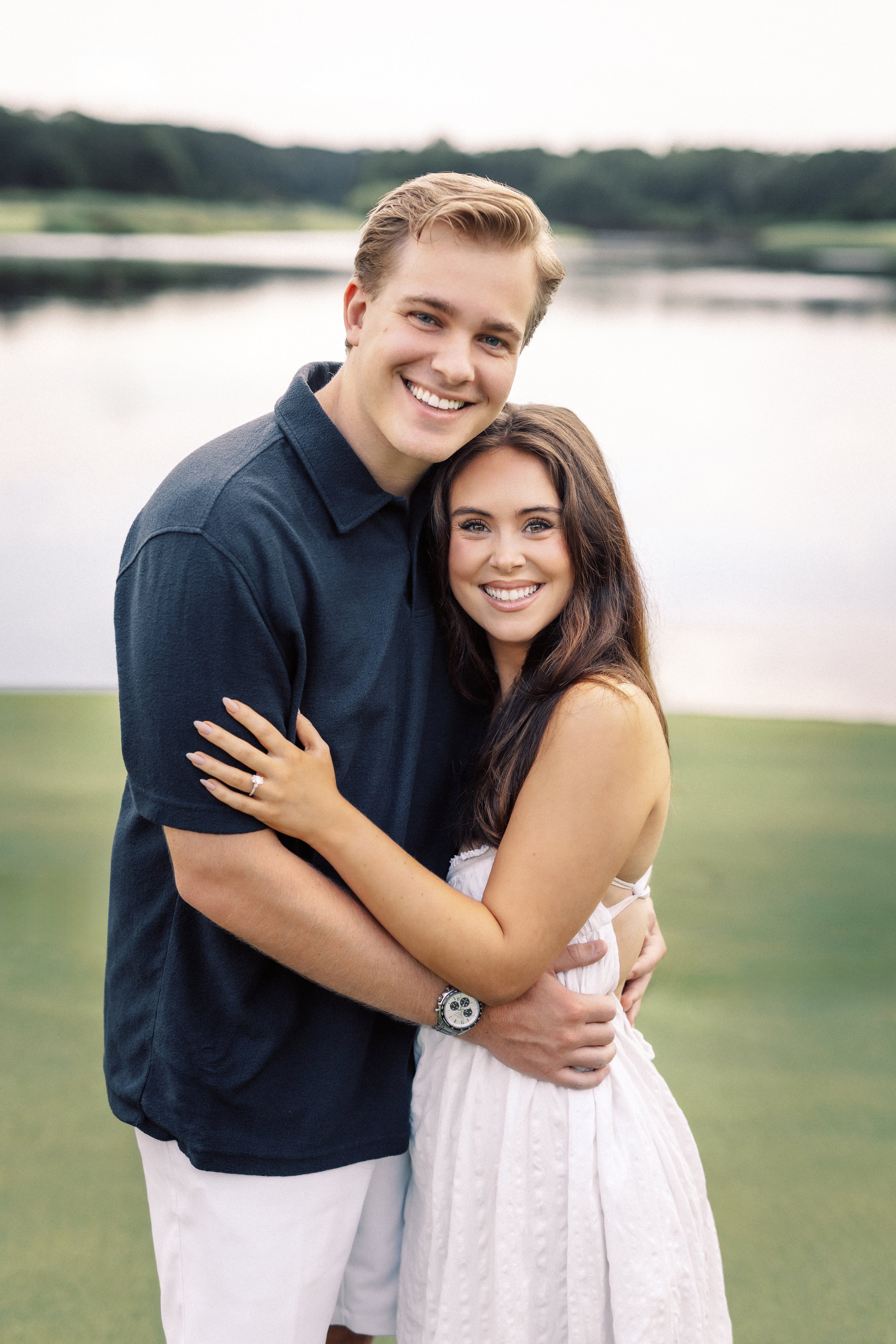 A happy couple embracing outdoors near a lake, with the man smiling broadly and the woman smiling softly, both looking at the camera.