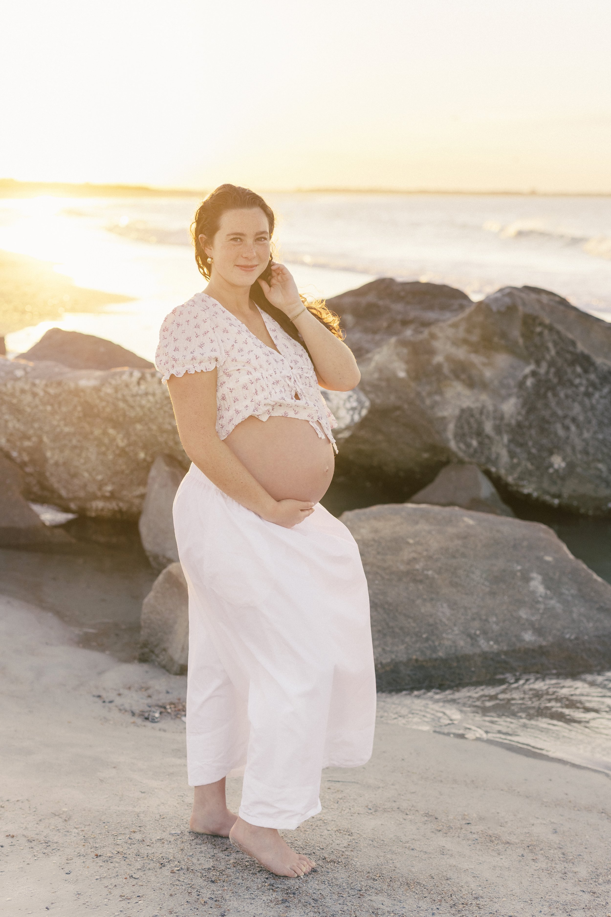 A pregnant woman in white pants and a light pink floral blouse standing barefoot on a sandy beach near large rocks, with the ocean and sunset in the background.