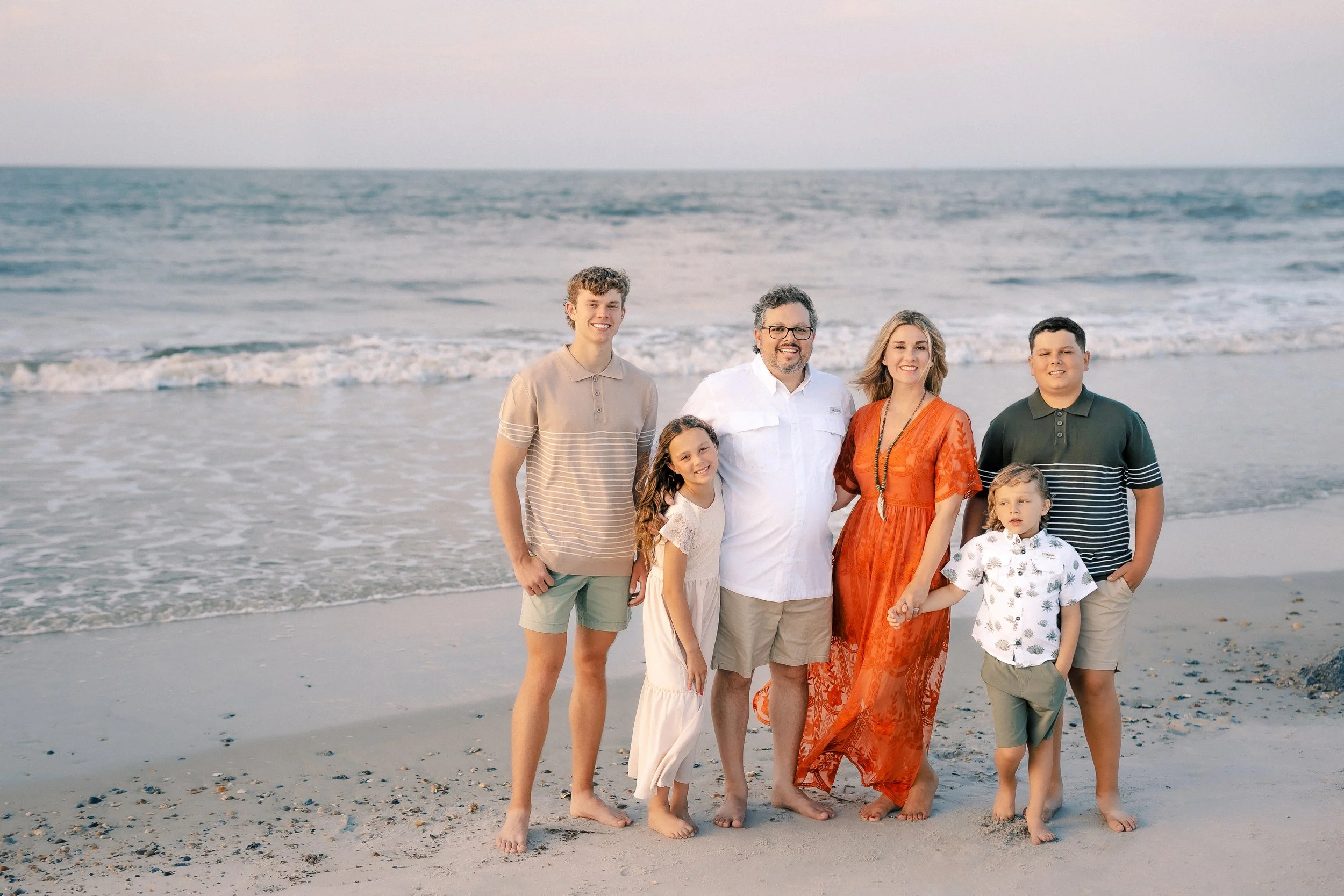 A family of eight standing on a beach near the shoreline with the ocean in the background, during sunset with soft lighting.
