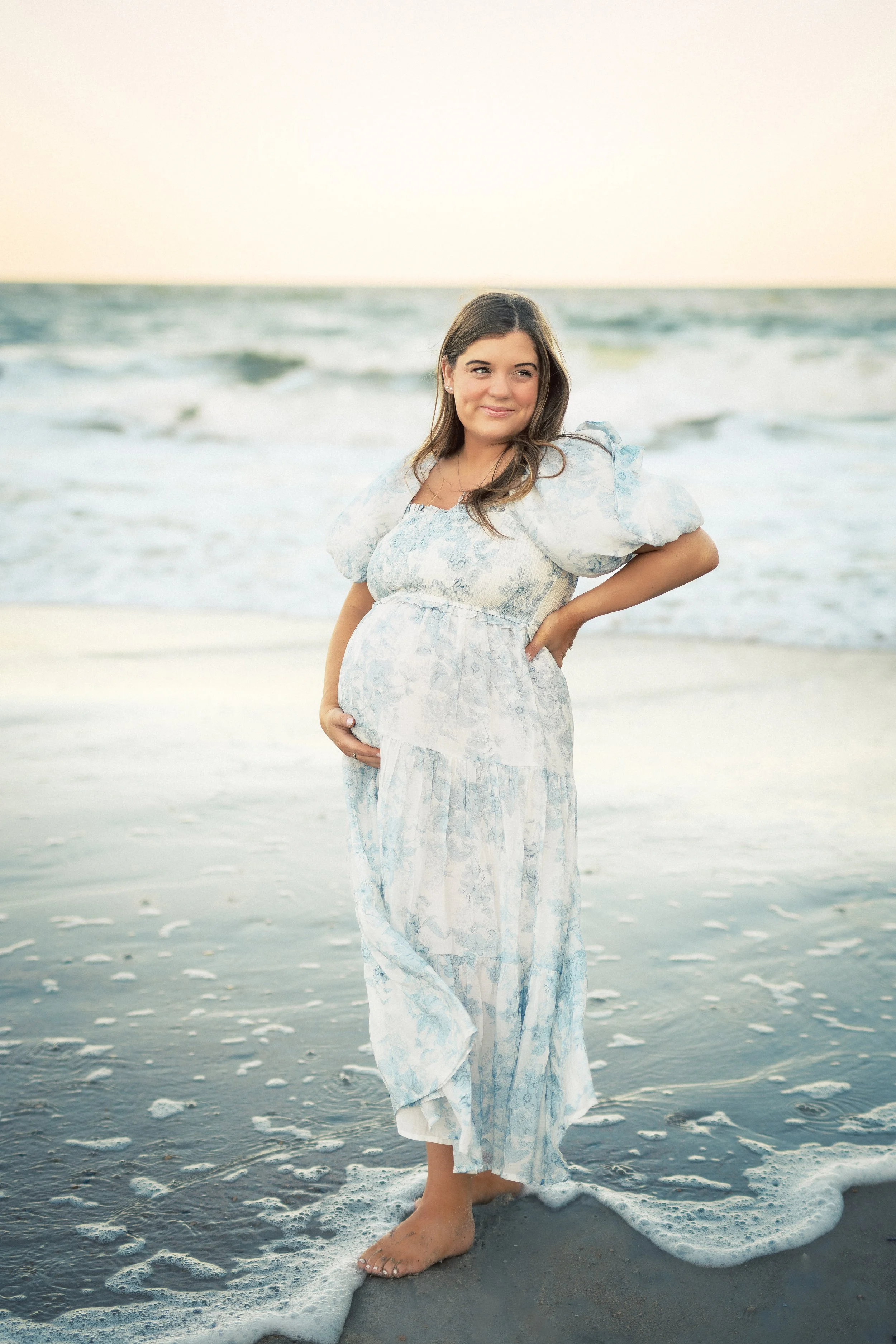 A woman in a light blue and white sundress standing on the beach, barefoot, with her hand on her hip, smiling slightly, ocean waves in the background at sunset.