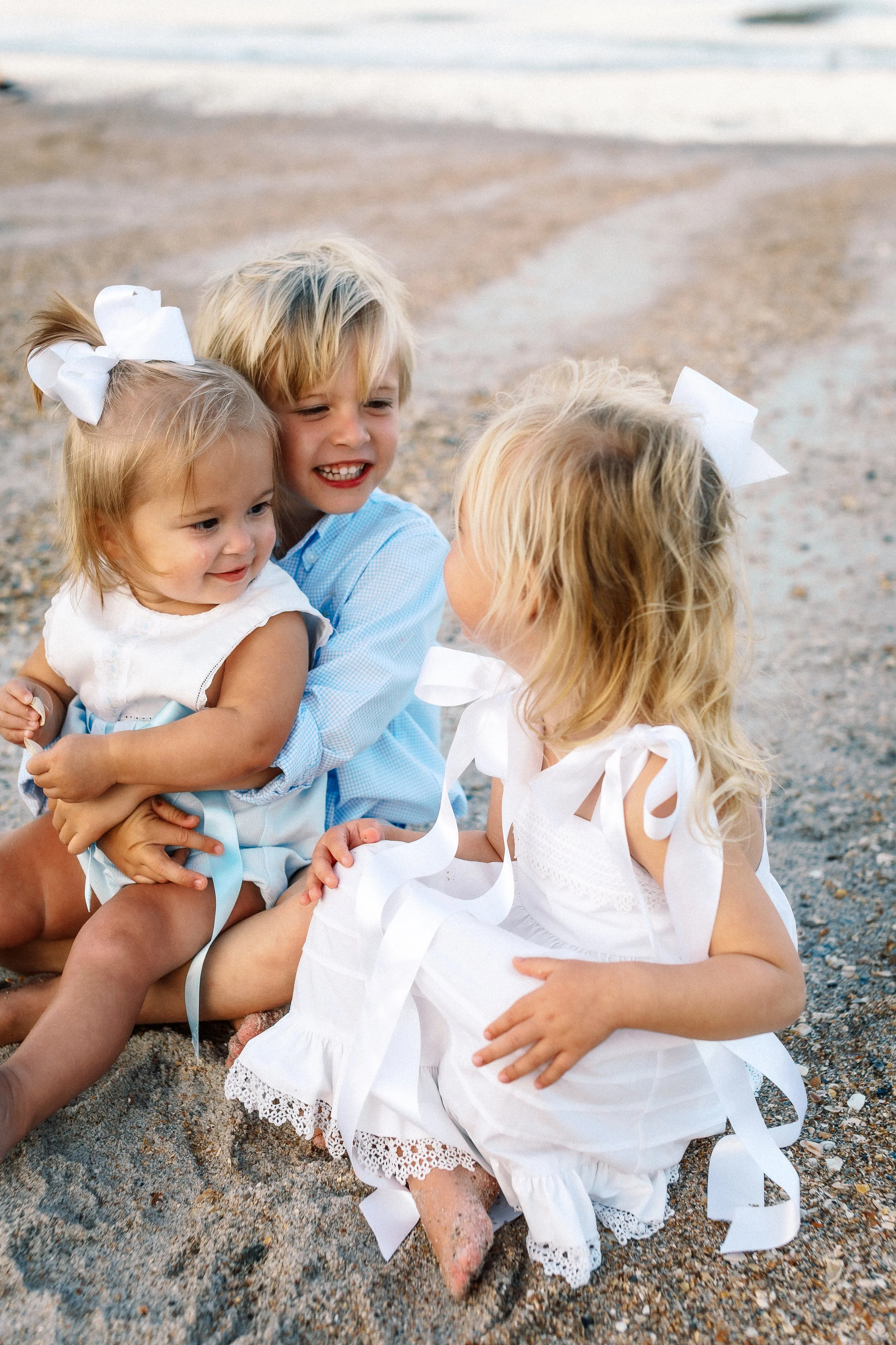 Four children playing on the beach, dressed in white dresses with bows in their hair, sitting on the sand near the water.Photographer Amelia Island, Jacksonville, St Augustine, St Simons Island, Savannah photographer