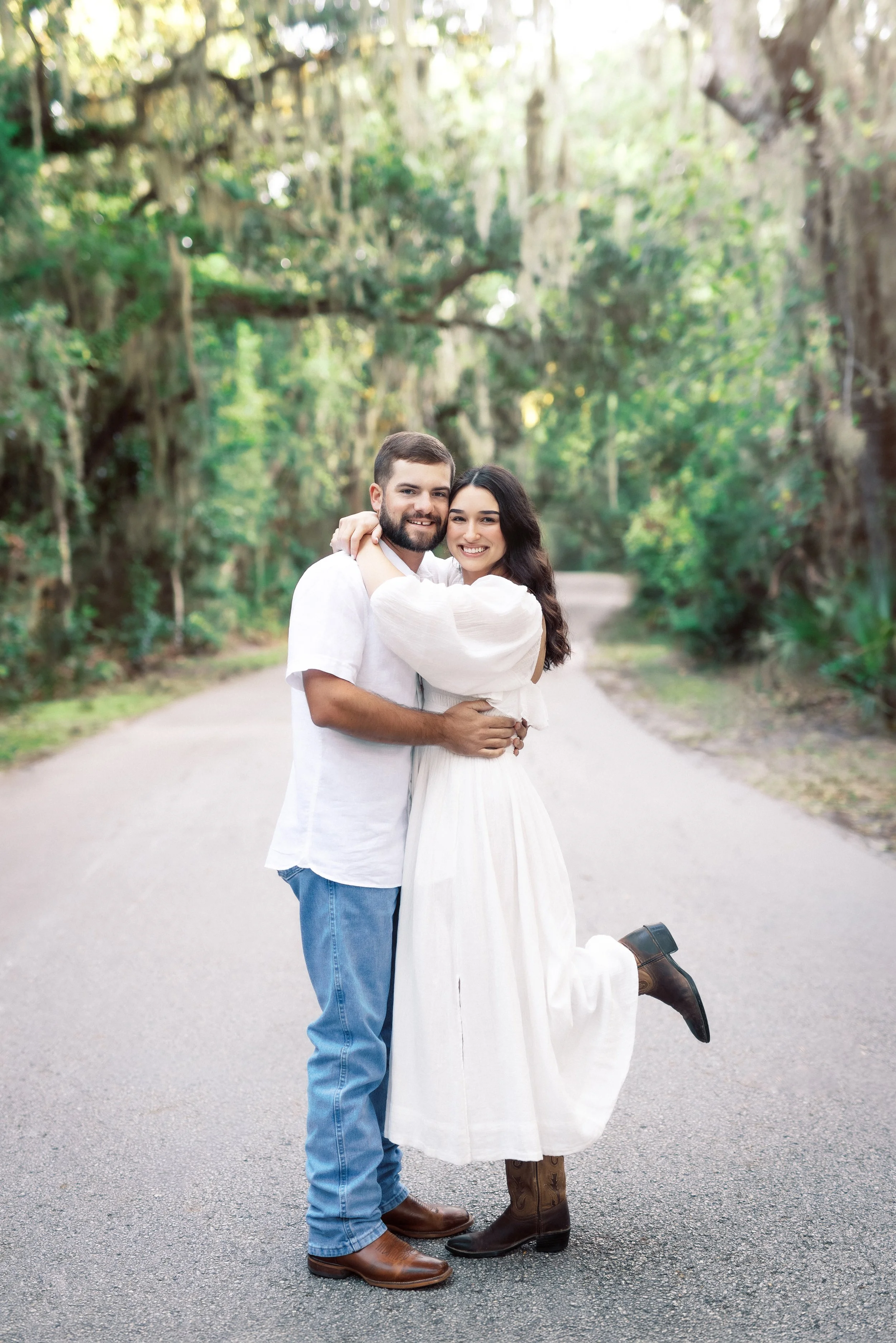 A happy couple stands in the middle of a tree-lined road, embracing and smiling at the camera. The woman, wearing a white dress and cowboy boots, is being lifted by the man, who is dressed casually in jeans and a white shirt. The sunlight filters thr