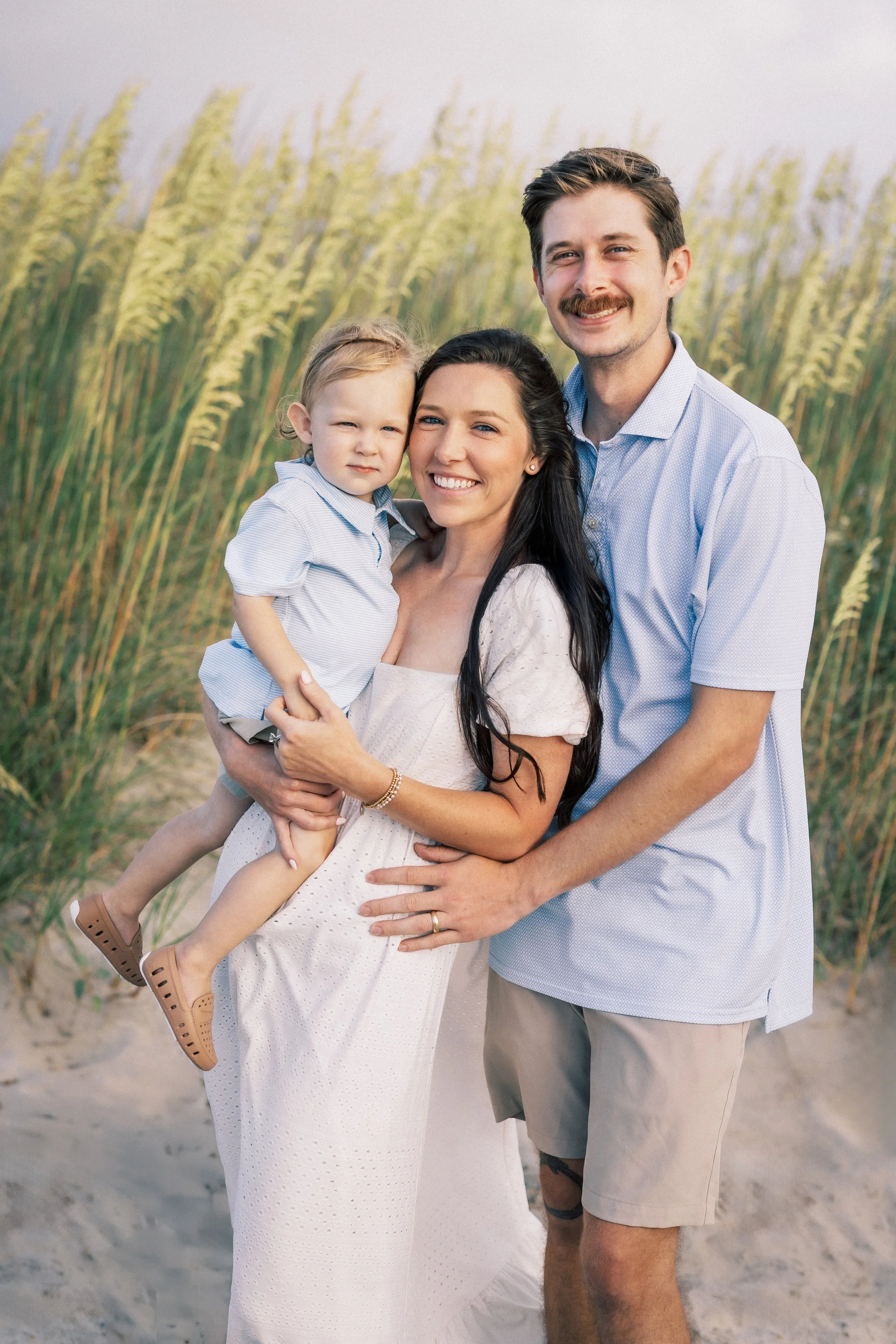 A happy family of three standing on a sandy beach with tall grasses in the background. The woman is holding a young girl, and the man is standing next to them with his arm around the woman. Photographer Amelia Island, Jacksonville, St Augustine, St S