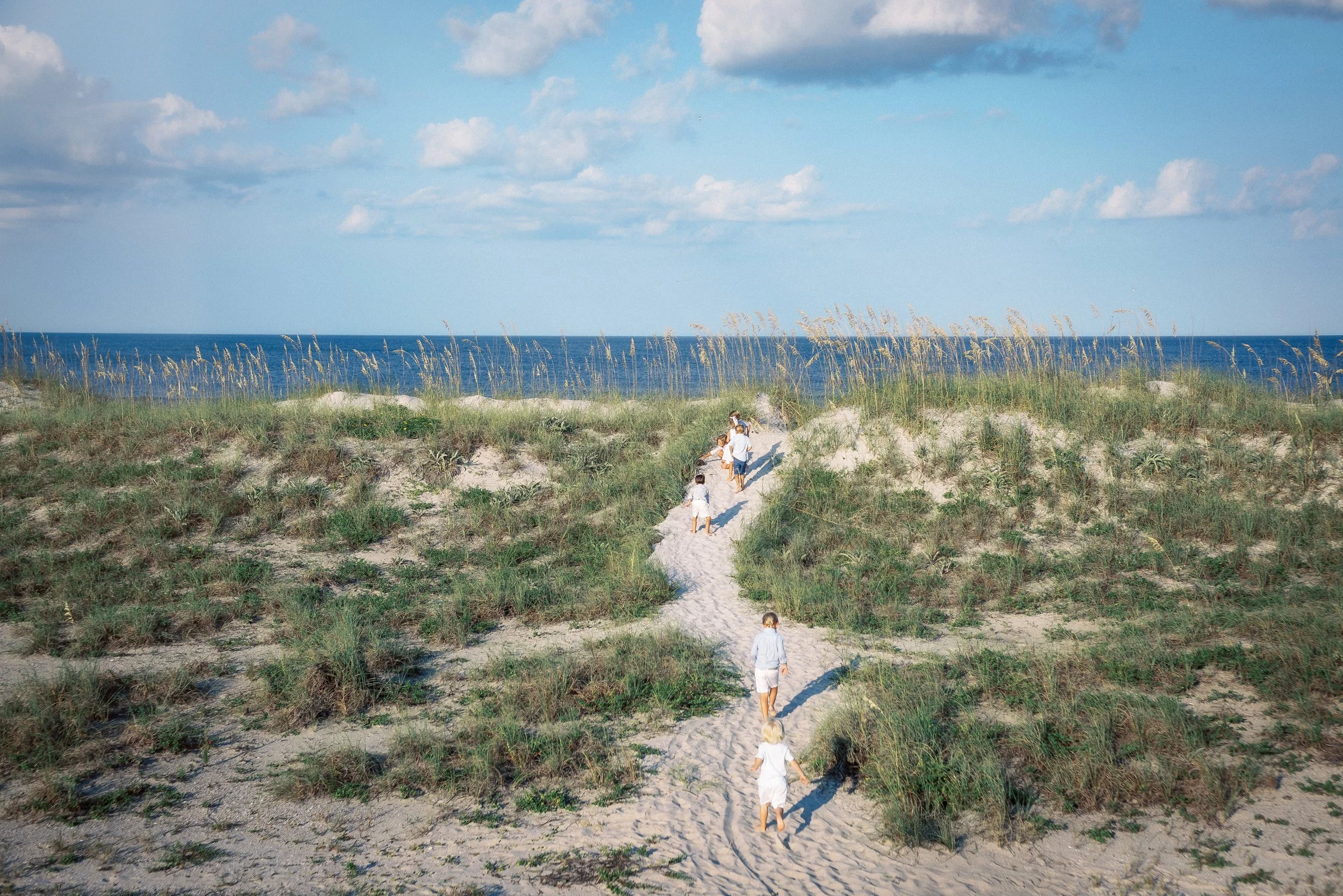 People walking along a sandy beach trail through grassy dunes with the ocean and blue sky with clouds in the background.Photographer Amelia Island, Jacksonville, St Augustine, St Simons Island, Savannah photographer