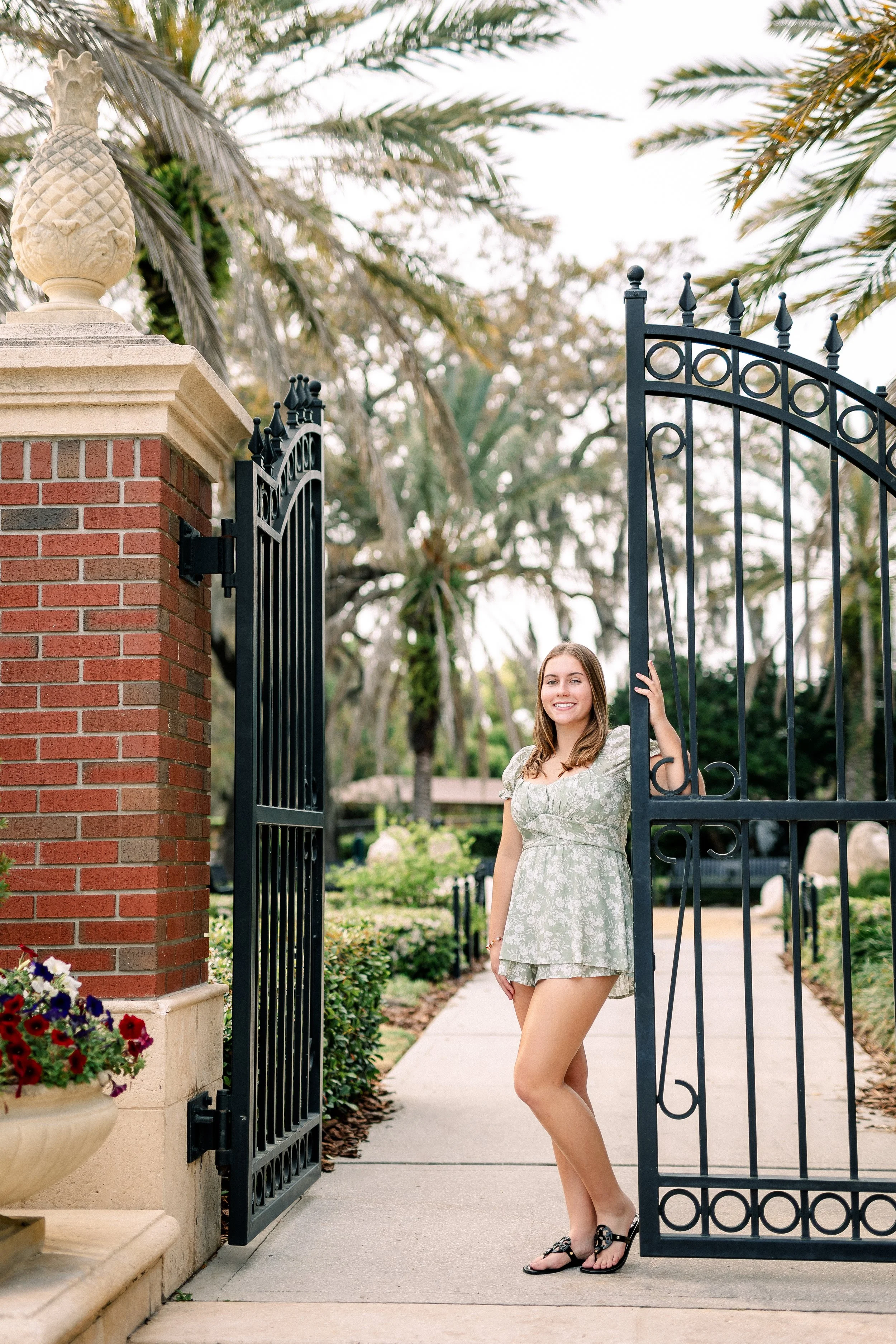 A young woman standing in a gate in a park or garden, smiling and looking at the camera. She is wearing a light green floral dress and sandals. The gate is black wrought iron, attached to a brick and stone pillar with a decorative finial. There are t