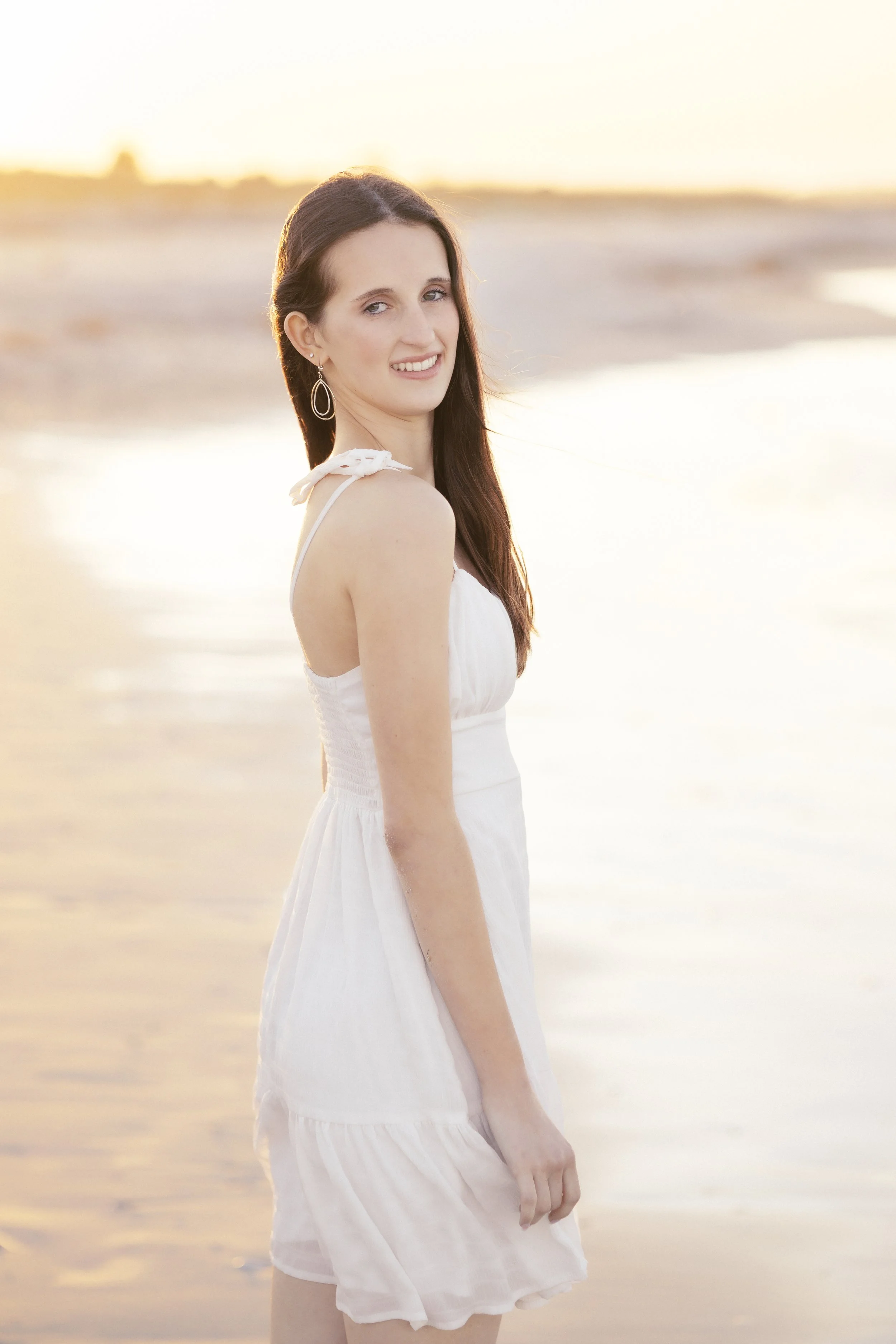 A woman in a white dress standing on the beach at sunset, smiling at the camera.
