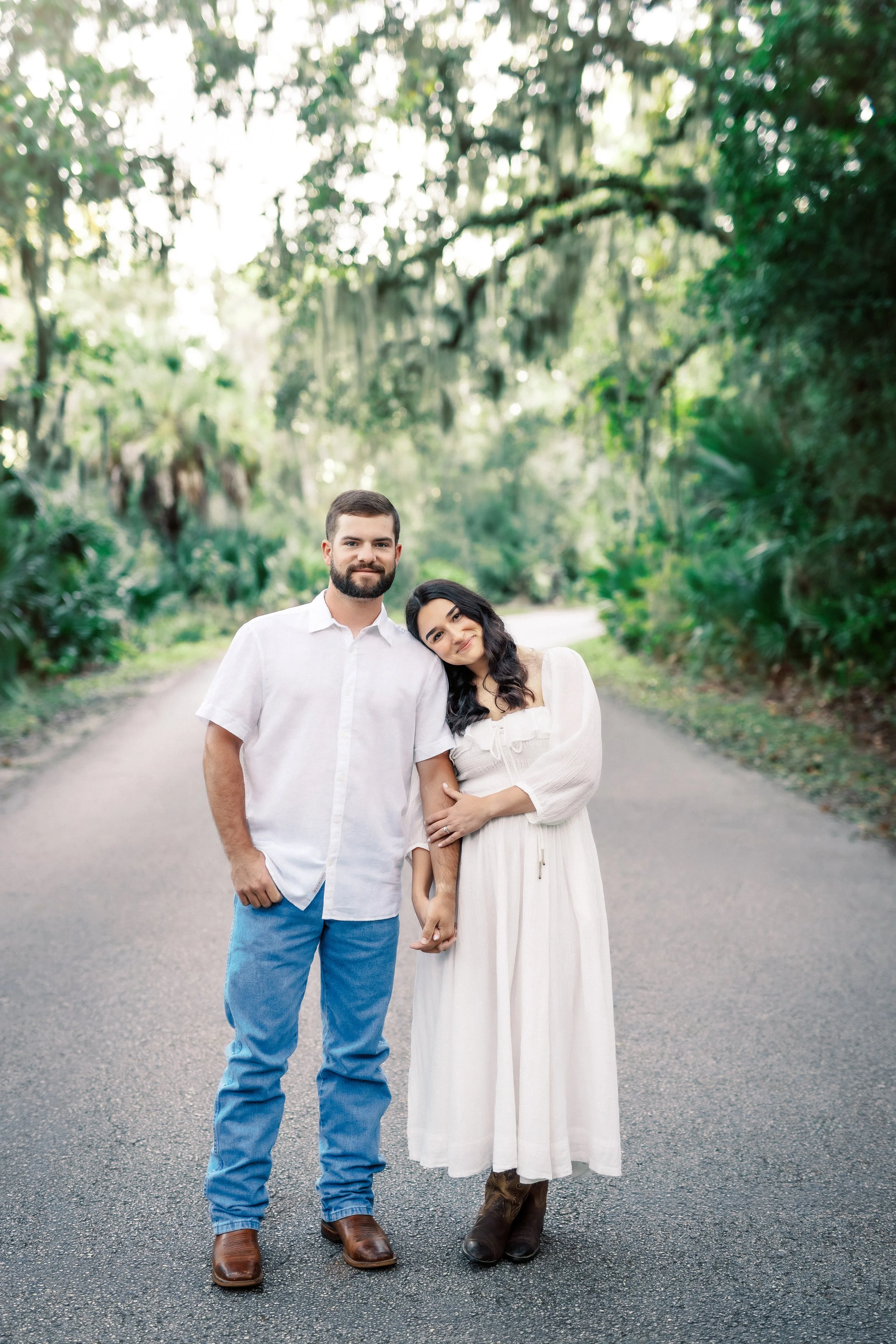 A couple standing hand in hand on a paved road in a lush, green wooded area. The man has a beard and is wearing a white shirt, blue pants, and brown boots. The woman has long dark hair and is wearing a white dress and brown boots, with her head resti