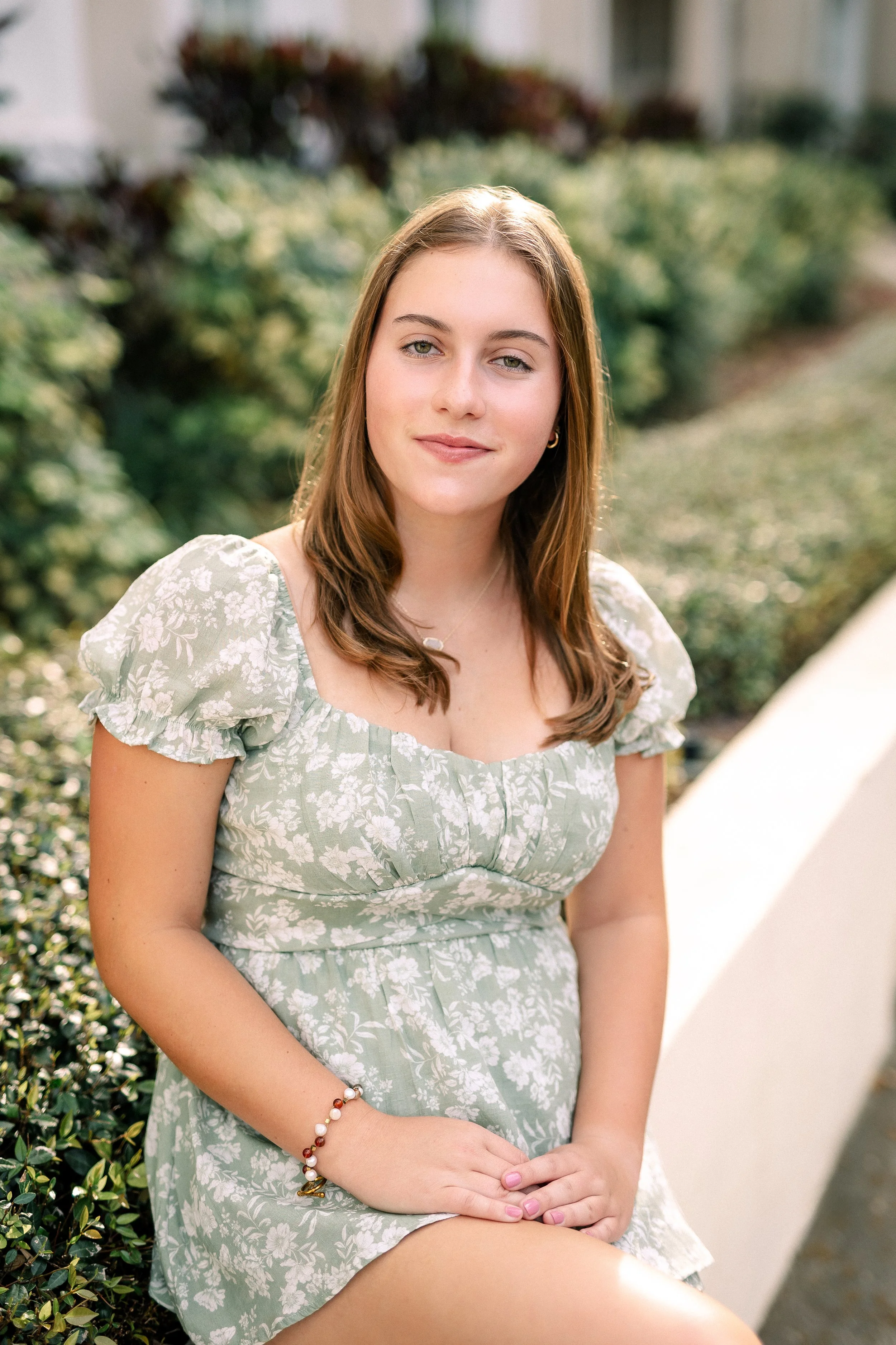 A young woman sitting outdoors with greenery behind her, wearing a light green floral dress and a beaded bracelet, looking into the camera with a slight smile.
